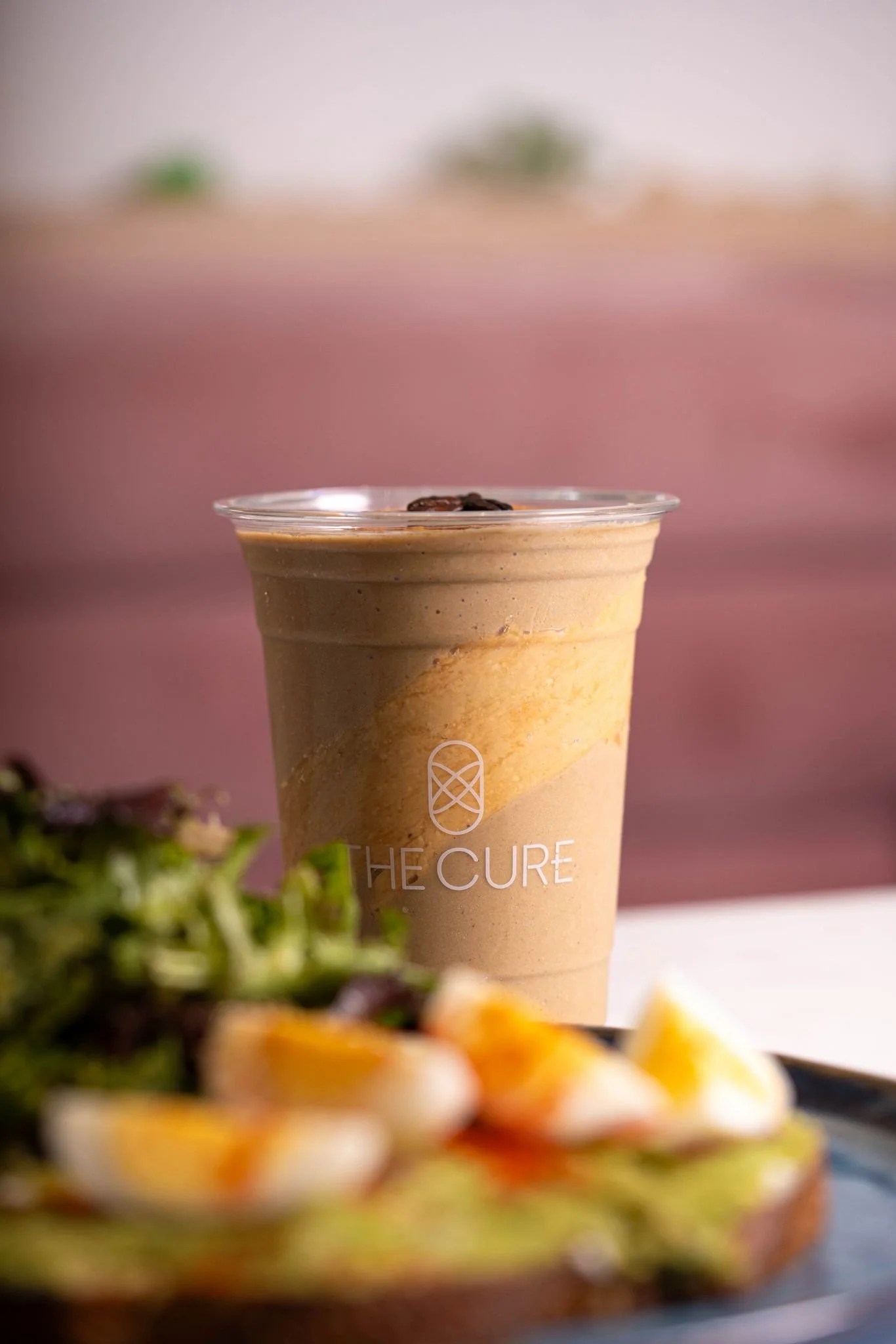 A cold coffee beverage in a clear plastic cup with 'The Cure' logo, placed on a white surface, with a salad and sliced boiled eggs in the foreground, blurred pink background.