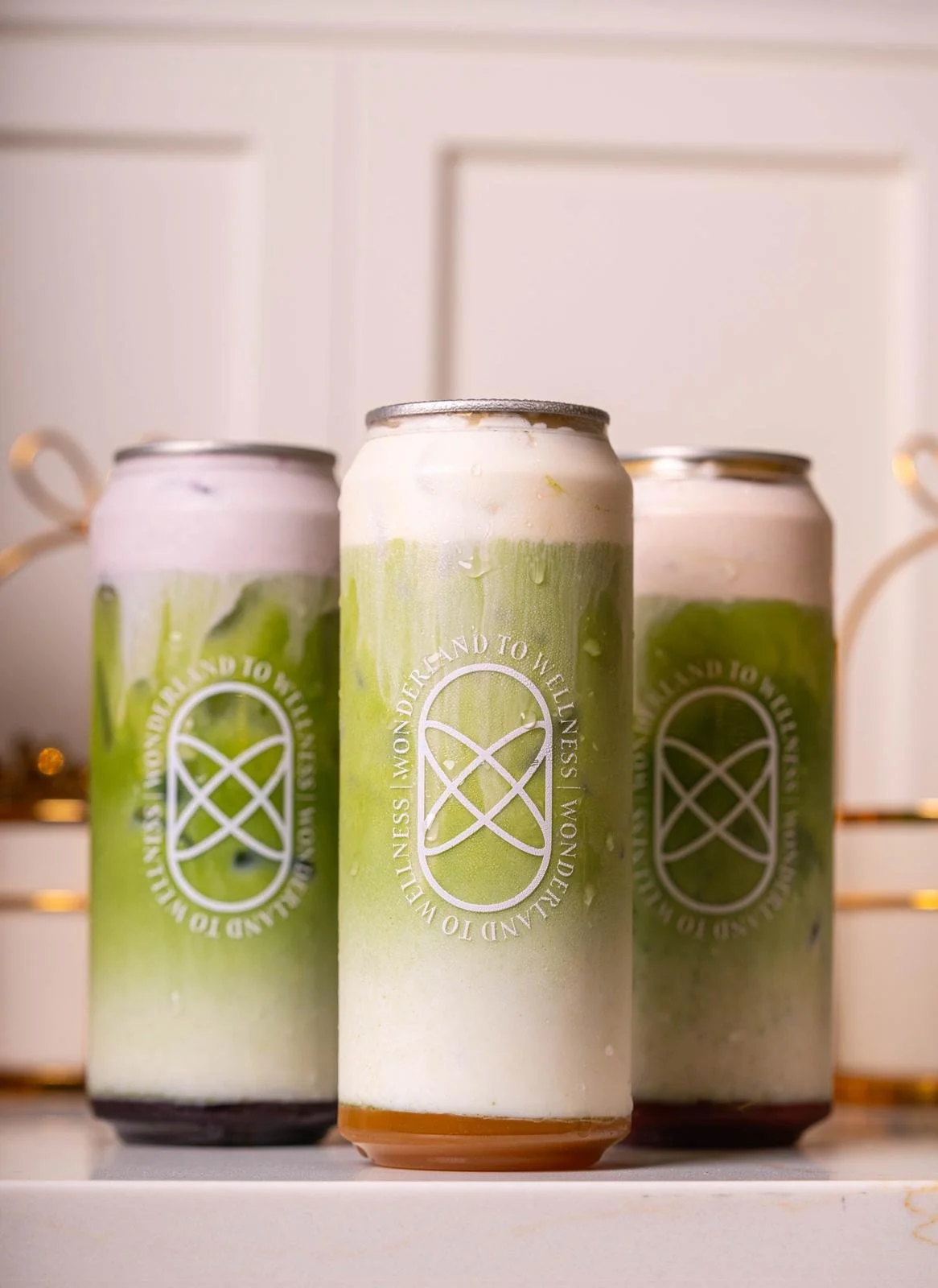Three cans of beverage with green and white labels, placed on a white surface with a white cabinet in the background.