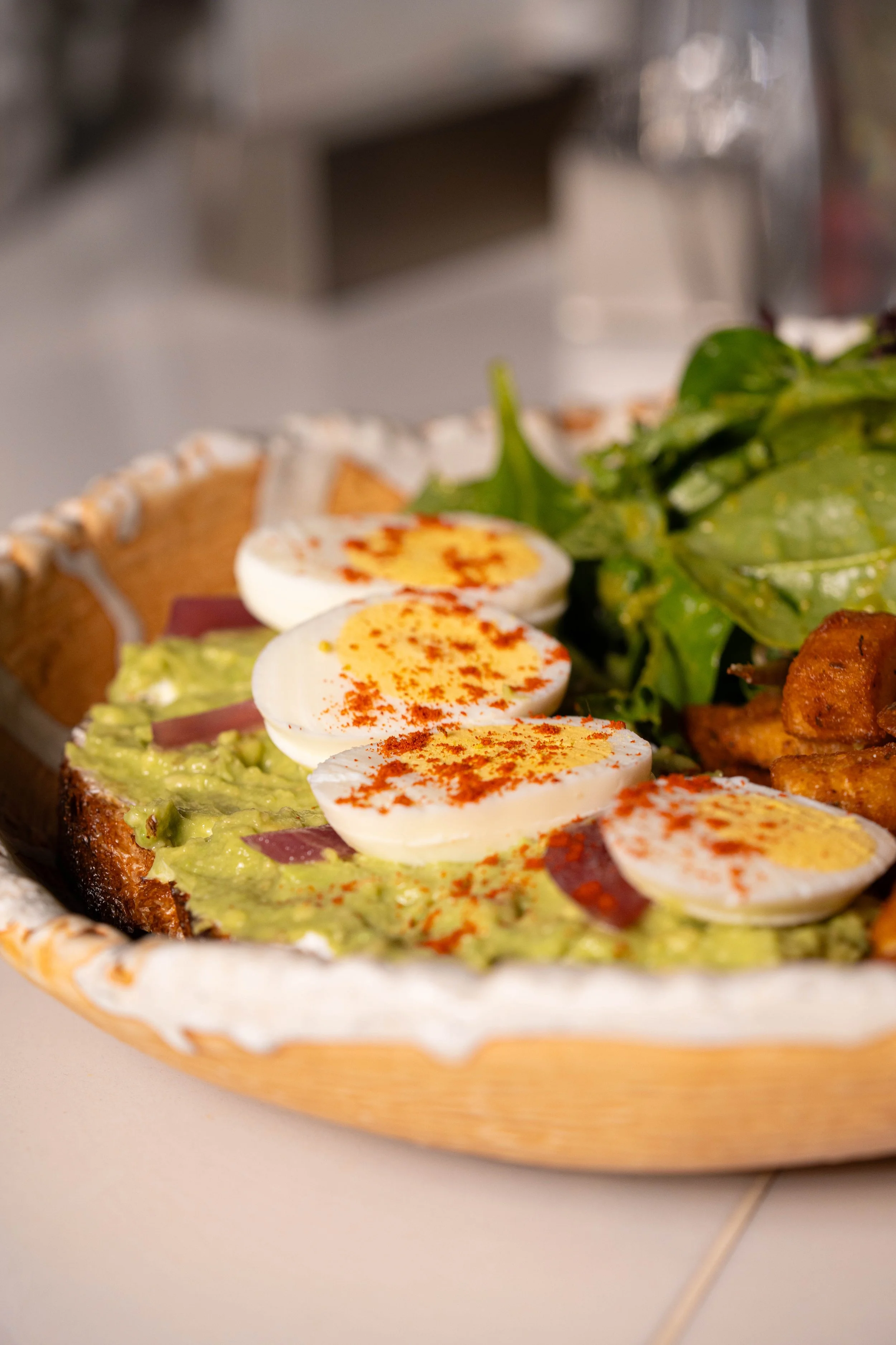 Close-up of a wooden platter with sliced avocado, boiled eggs sprinkled with paprika, bacon bits, mixed greens salad, and toast visible in the background.