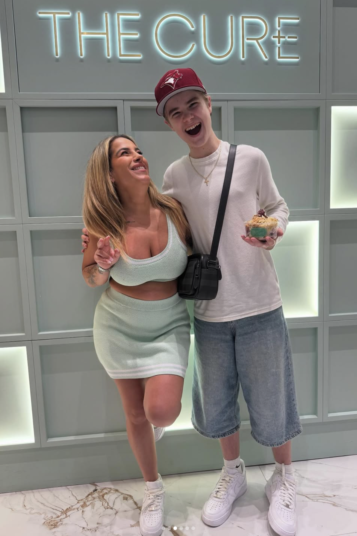 A woman and a young man are standing together in front of a sign that reads 'THE CURE'. The woman is smiling and wearing a light green crop top and matching skirt, while the young man is laughing and wearing a white long-sleeve shirt, jean shorts, and a red cap. The woman has her arm around the young man's waist, and he is holding a bowl of dessert.