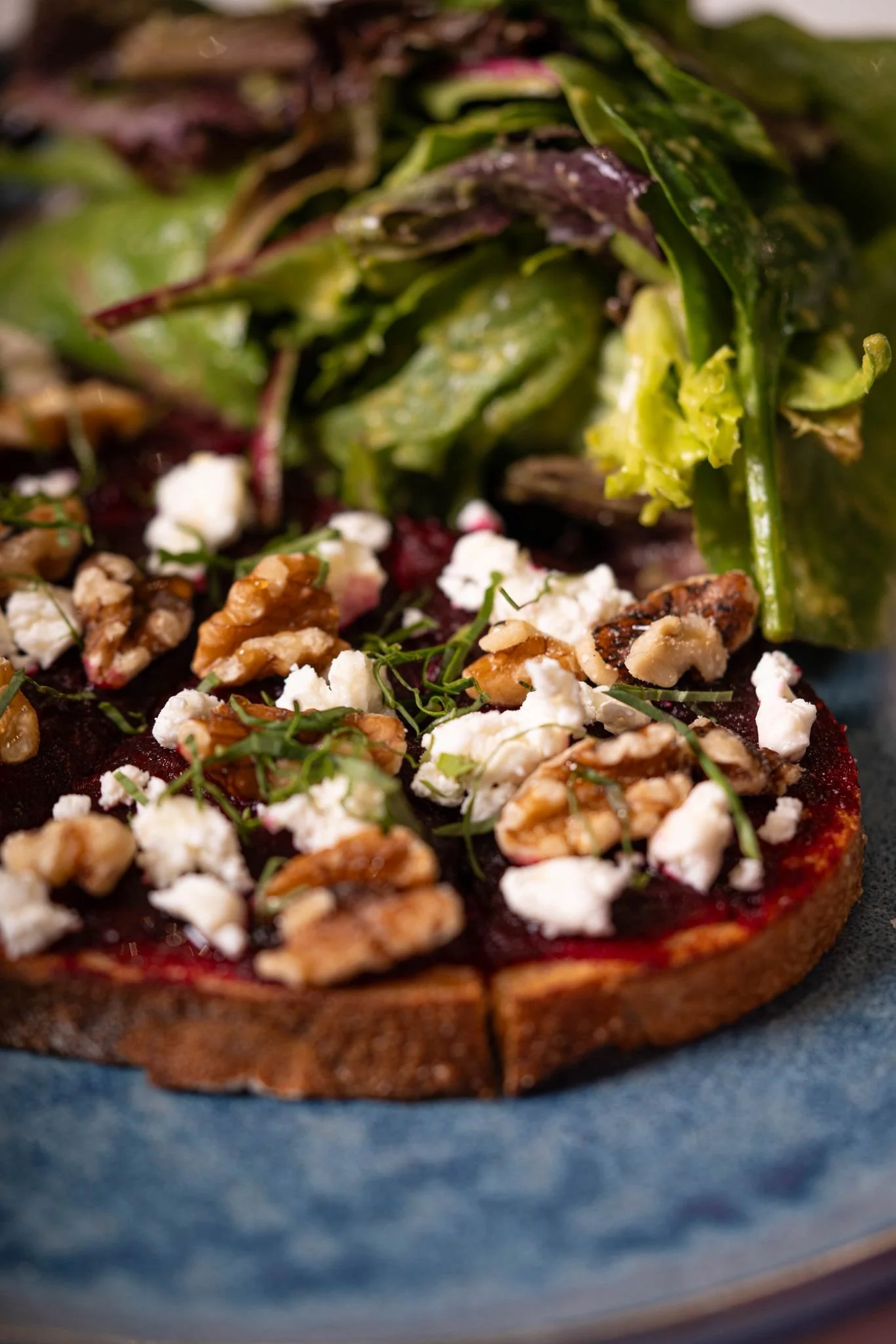 Close-up of a slice of beet tartare topped with crumbled cheese, walnut pieces, and fresh herbs, served with a side of mixed greens.