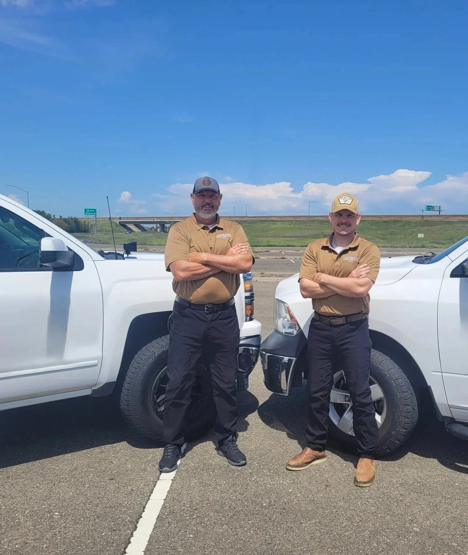 Two men in uniform standing between two white vehicles on a highway, with a blue sky and some clouds in the background.