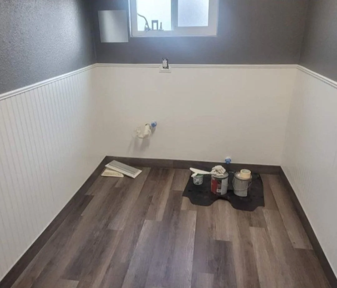 Room under renovation with unfinished flooring, wainscoting on walls, and painting supplies on the floor.