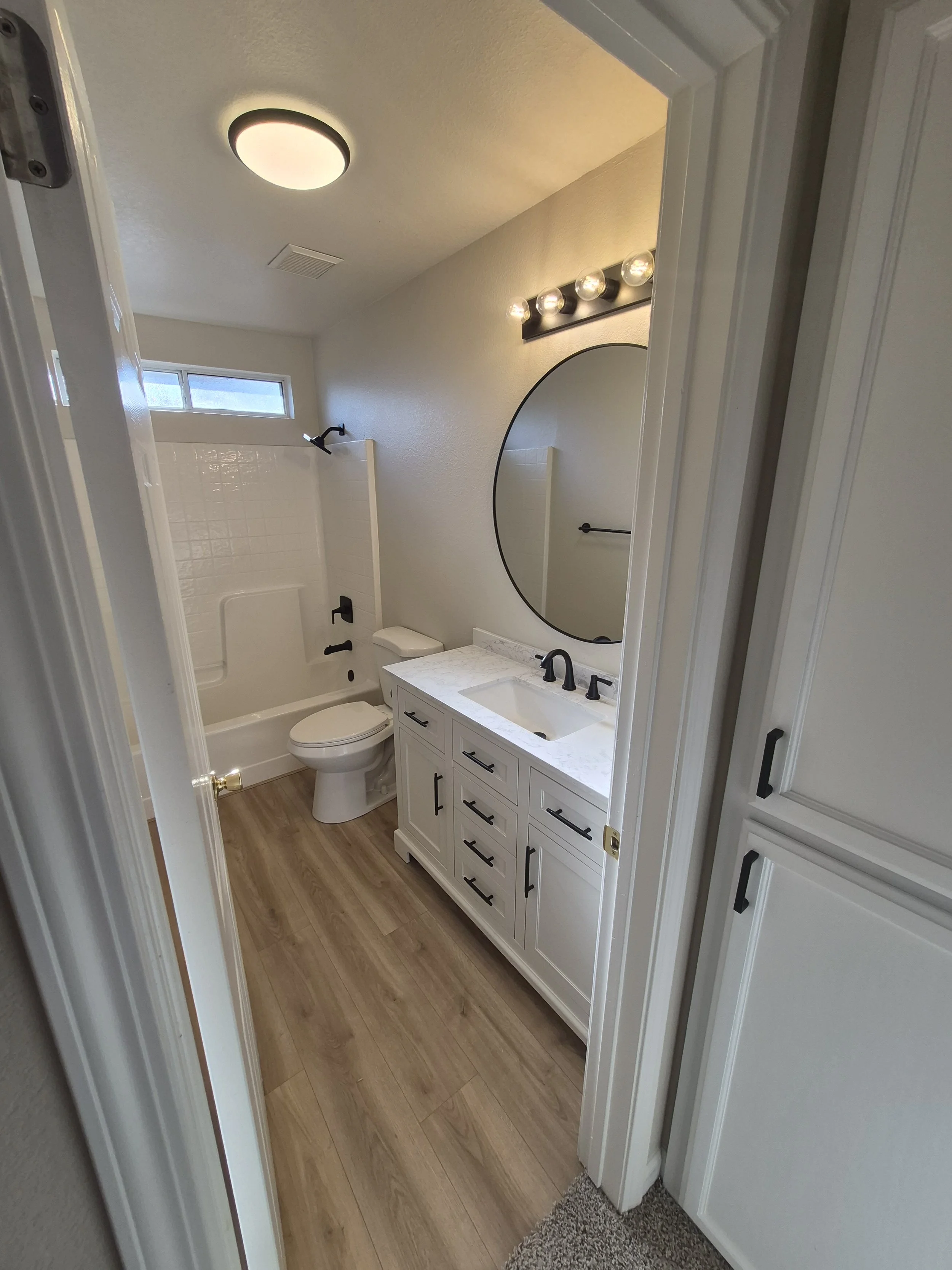 A bathroom with white cabinetry, a marble countertop, a round mirror, a toilet, a shower with black fixtures, a small window, and wood-look flooring.