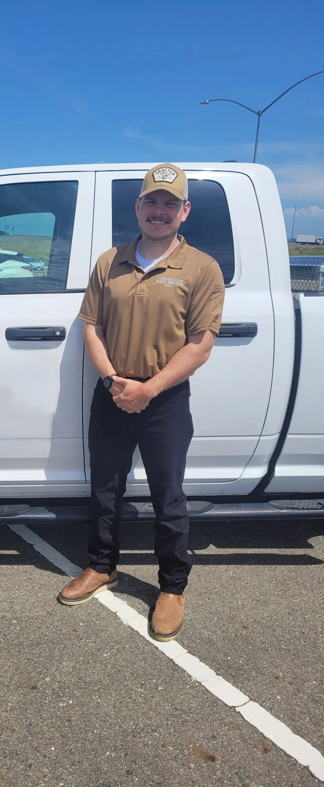 A man dressed in a tan work shirt and tan cap standing in front of a white pickup truck in a parking lot under a blue sky.