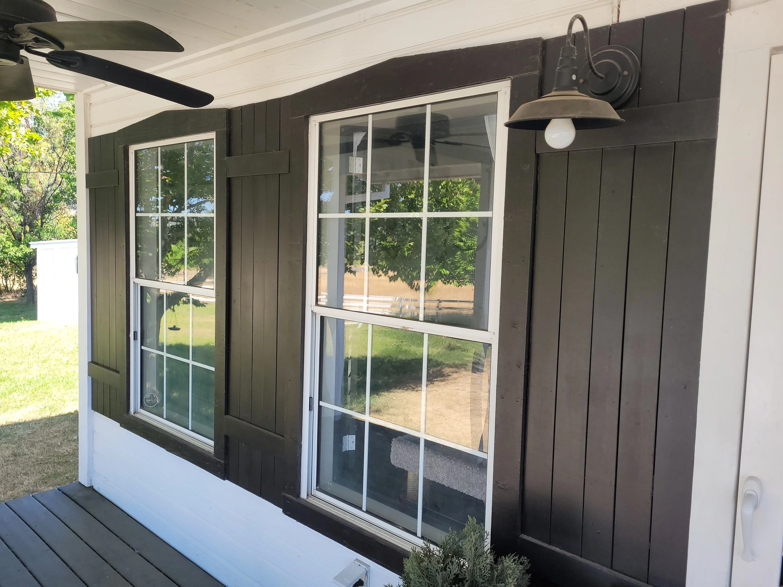 Exterior view of a house with dark wooden siding, two large windows, a black wall-mounted outdoor light, and a ceiling fan on the porch, with green trees and grass in the background.