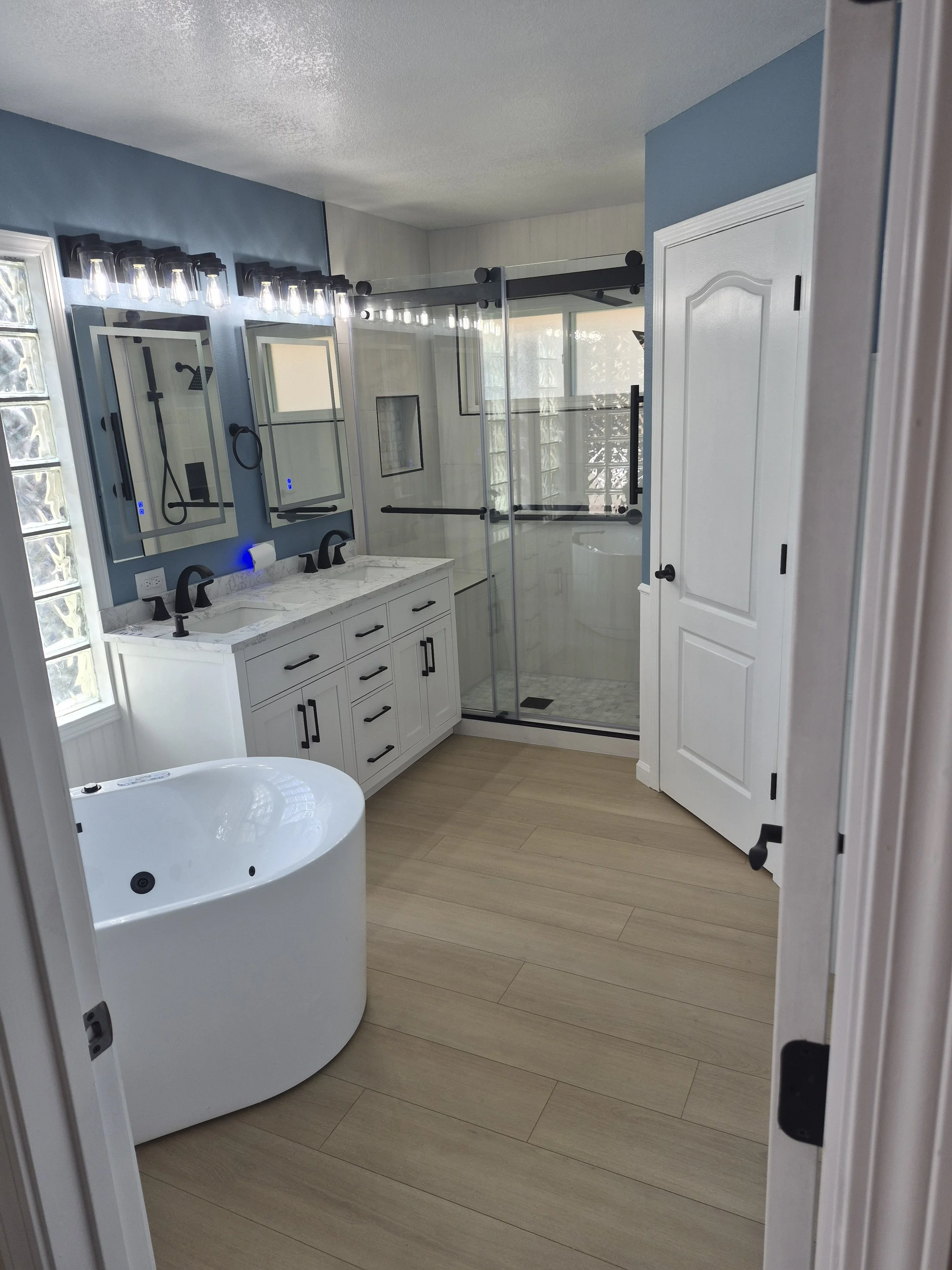 Bathroom with a walk-in shower with glass sliding doors, dual vanity with marble countertop and black fixtures, and a white freestanding bathtub. Light wood flooring, blue and white walls, and LED vanity lighting.