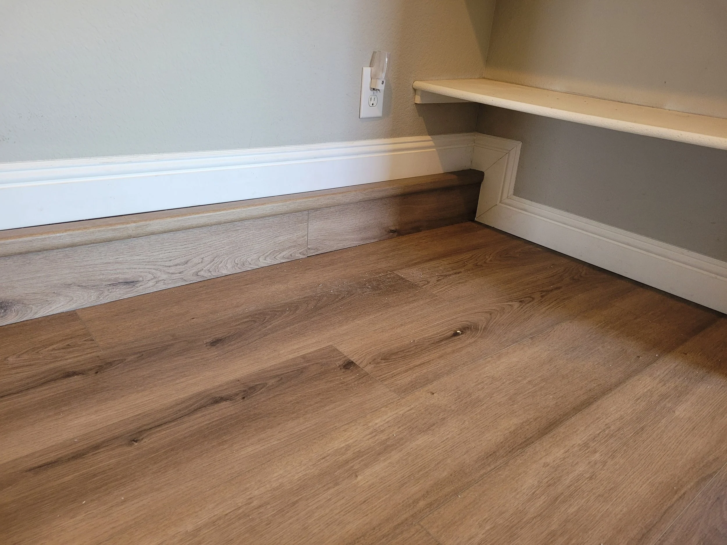 Wooden floor with dust and debris, baseboards, electrical outlet, and a wall with a corner shelf.