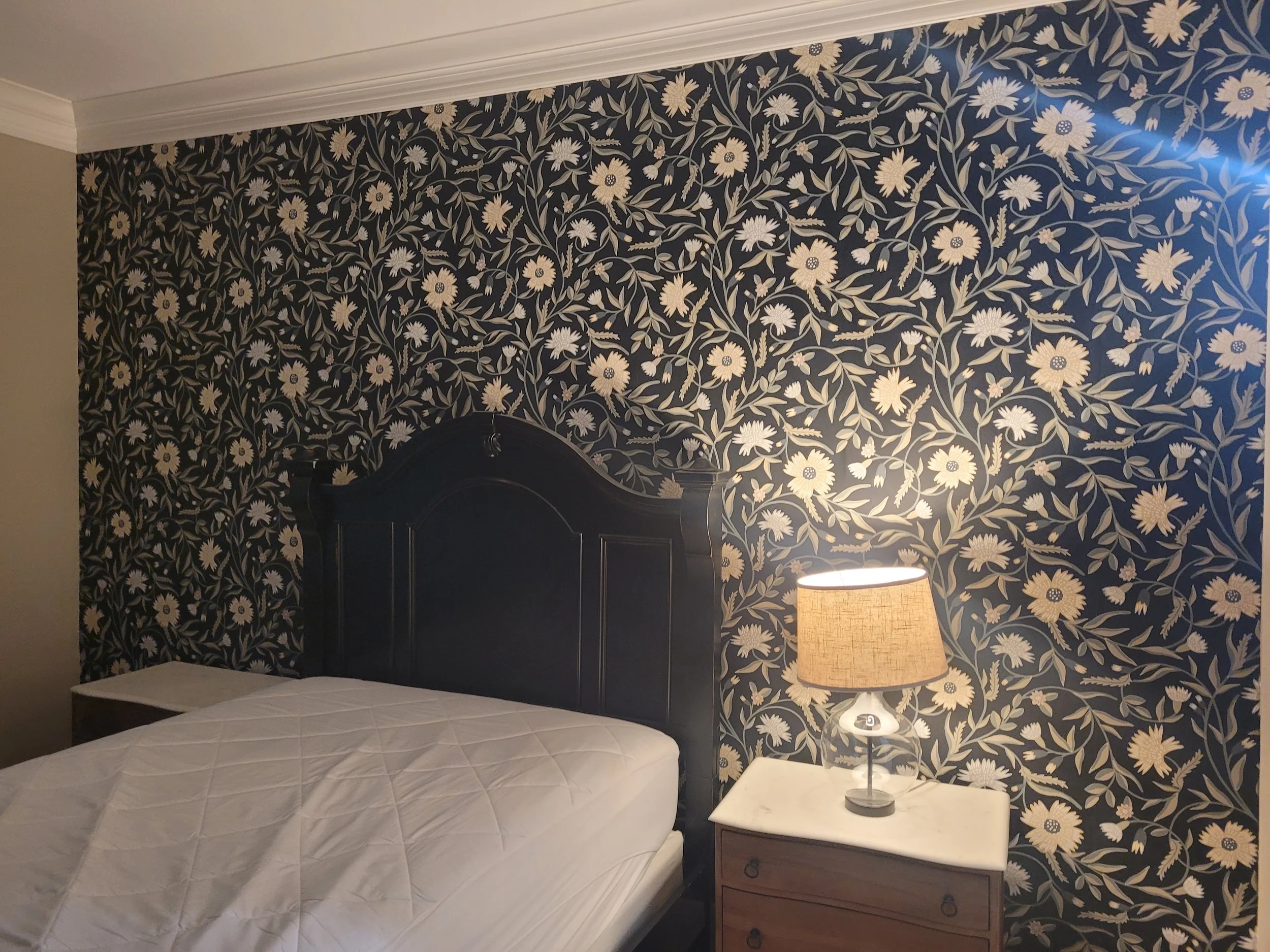 A bedroom with a dark wooden bed headboard, floral wallpaper, a white table lamp with a beige shade on a side table, and a white bedspread.