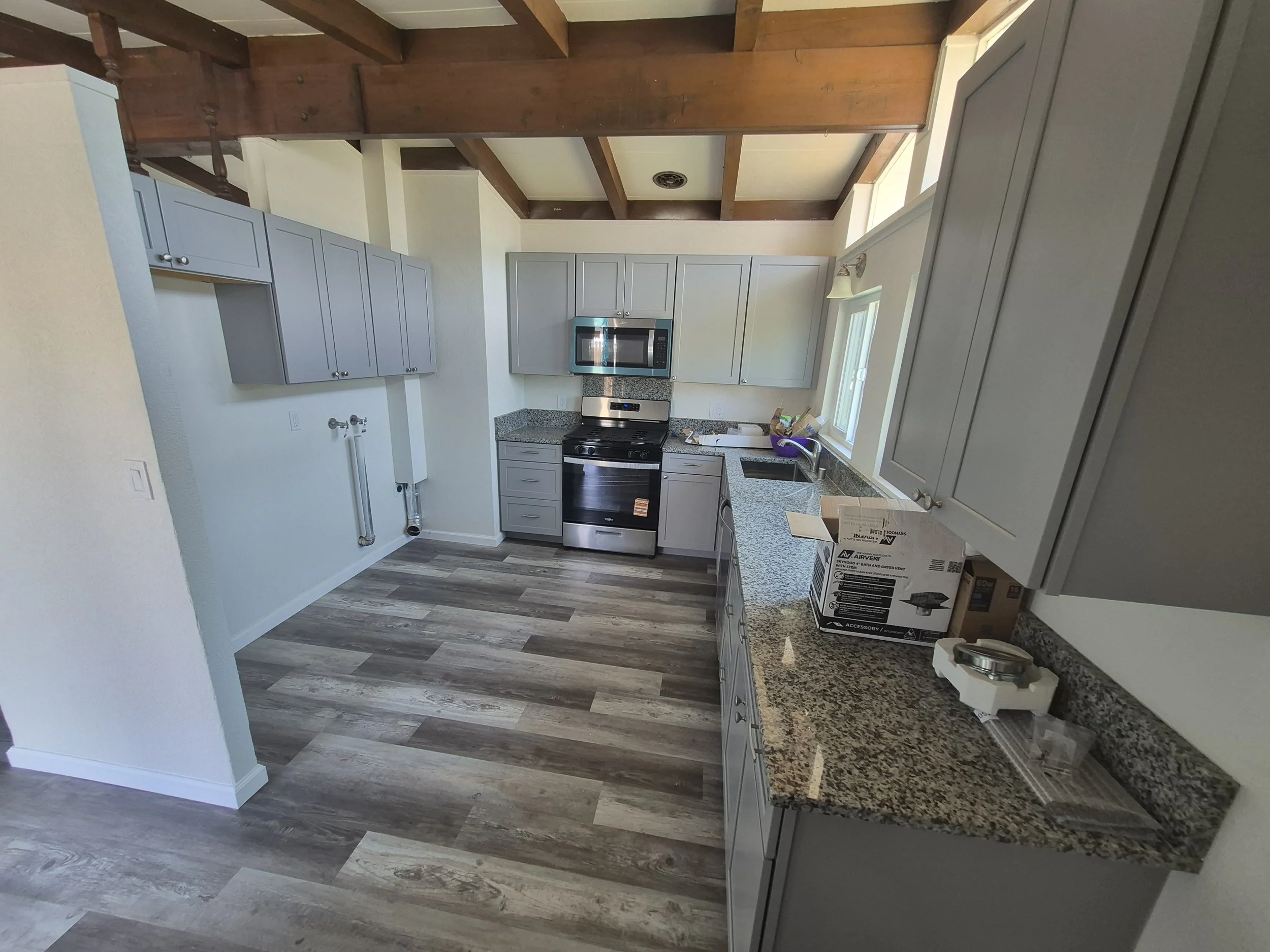 A modern kitchen with gray cabinets, stainless steel appliances, granite countertops, and hardwood flooring. The view includes a window, a microwave, a stove, and some boxes on the counter.