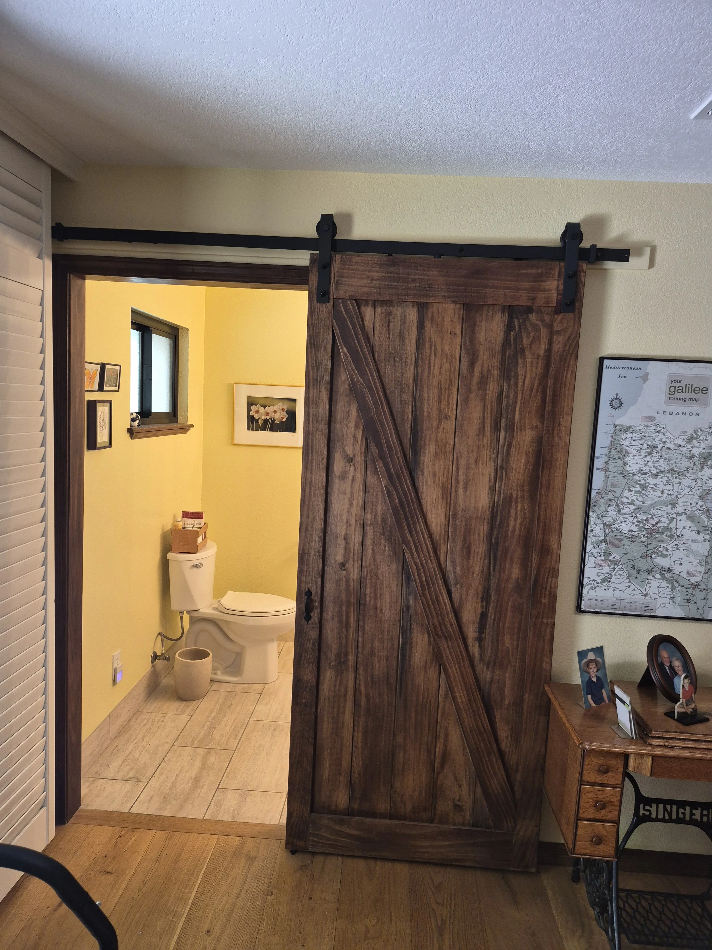 A sliding barn door partially open to reveal a bathroom with a toilet, framed artwork, and a small window.