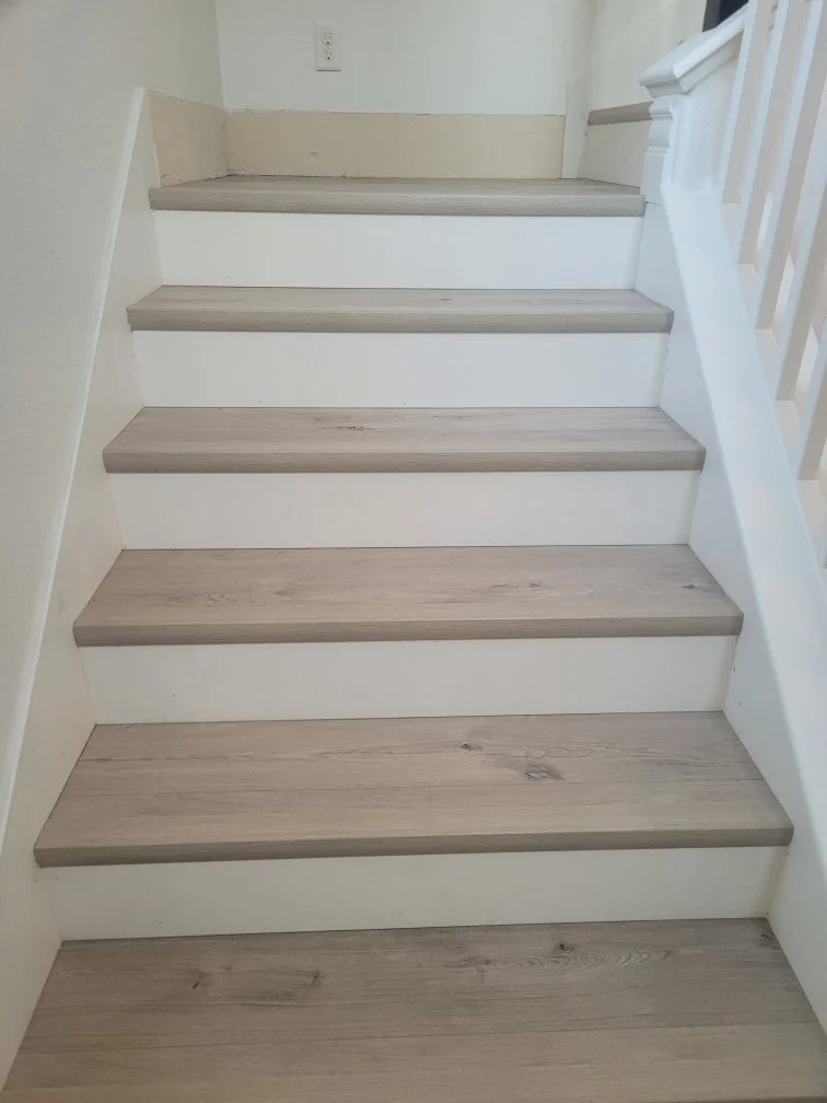 A staircase with light wood steps and white risers, adjacent to a white wall with a black outlet near the top, and a white balustrade on the right side.