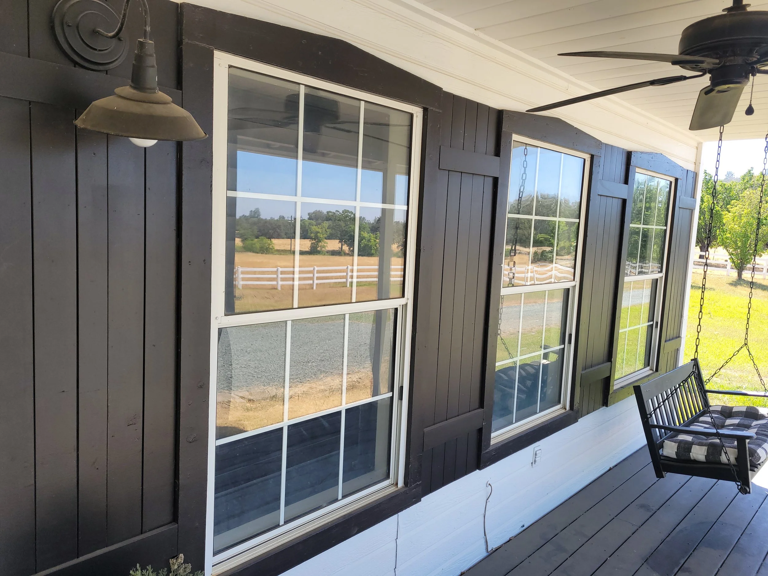 View of a house porch with three large windows, black exterior siding, a porch swing with plaid cushion, outdoor ceiling fan, and an outdoor wall light, overlooking a farm with fields and trees.