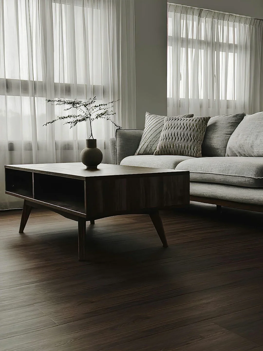 Living room with a grey sofa, patterned cushions, a wooden coffee table with a vase and branch, and sheer curtains in the background.