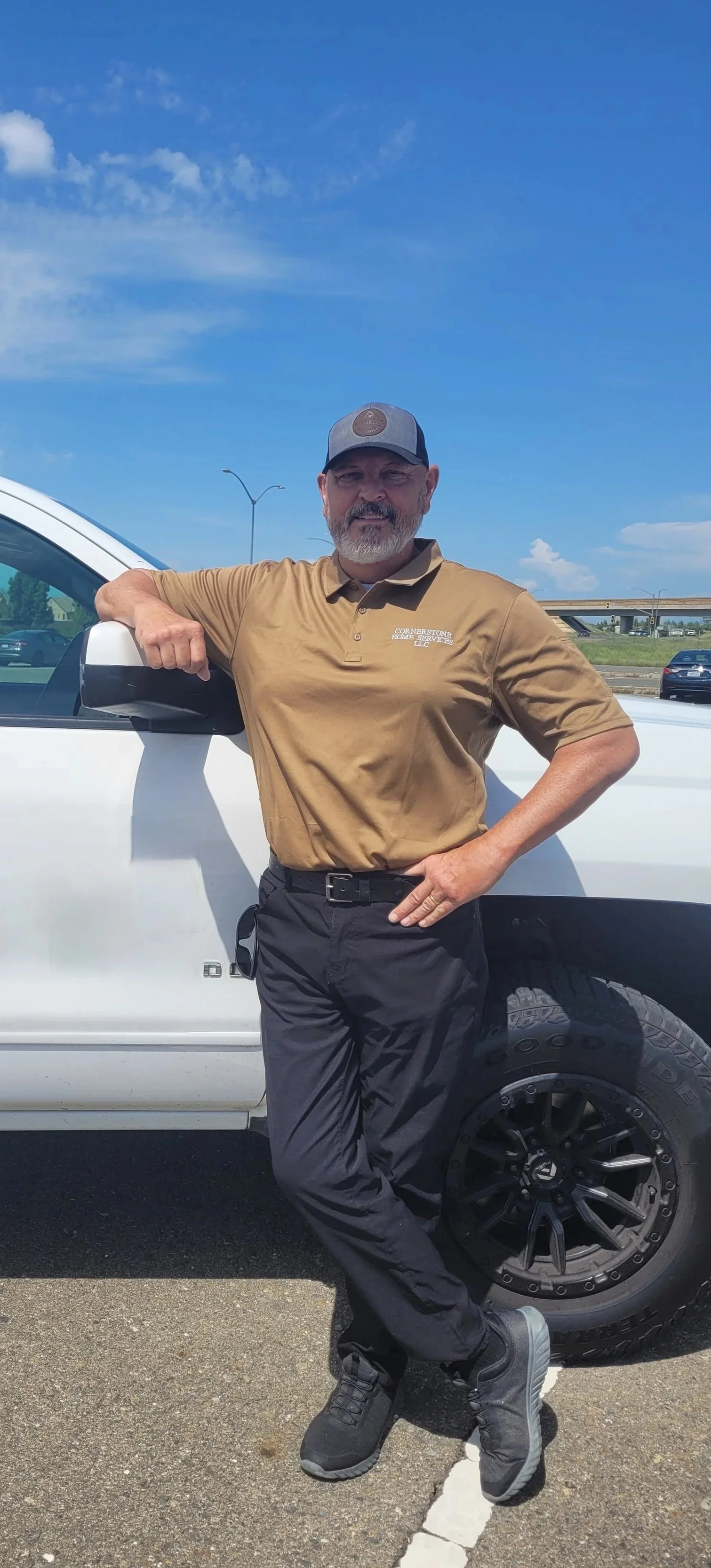 A man in a tan polo shirt and a cap standing next to a white pickup truck on a parking lot, with blue sky and some clouds in the background.