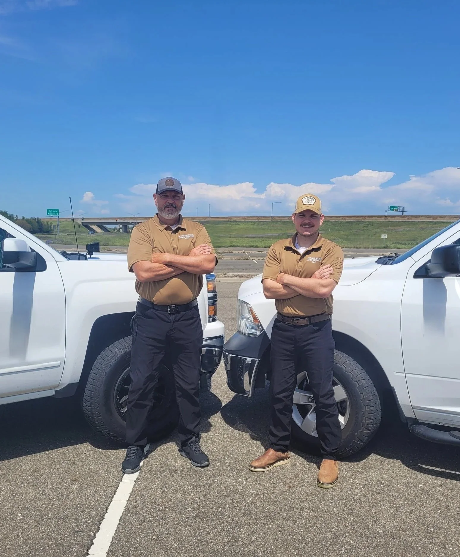 Two men in tan shirts and black pants standing in front of white trucks on a highway, with a grassy slope and clear blue sky in the background.
