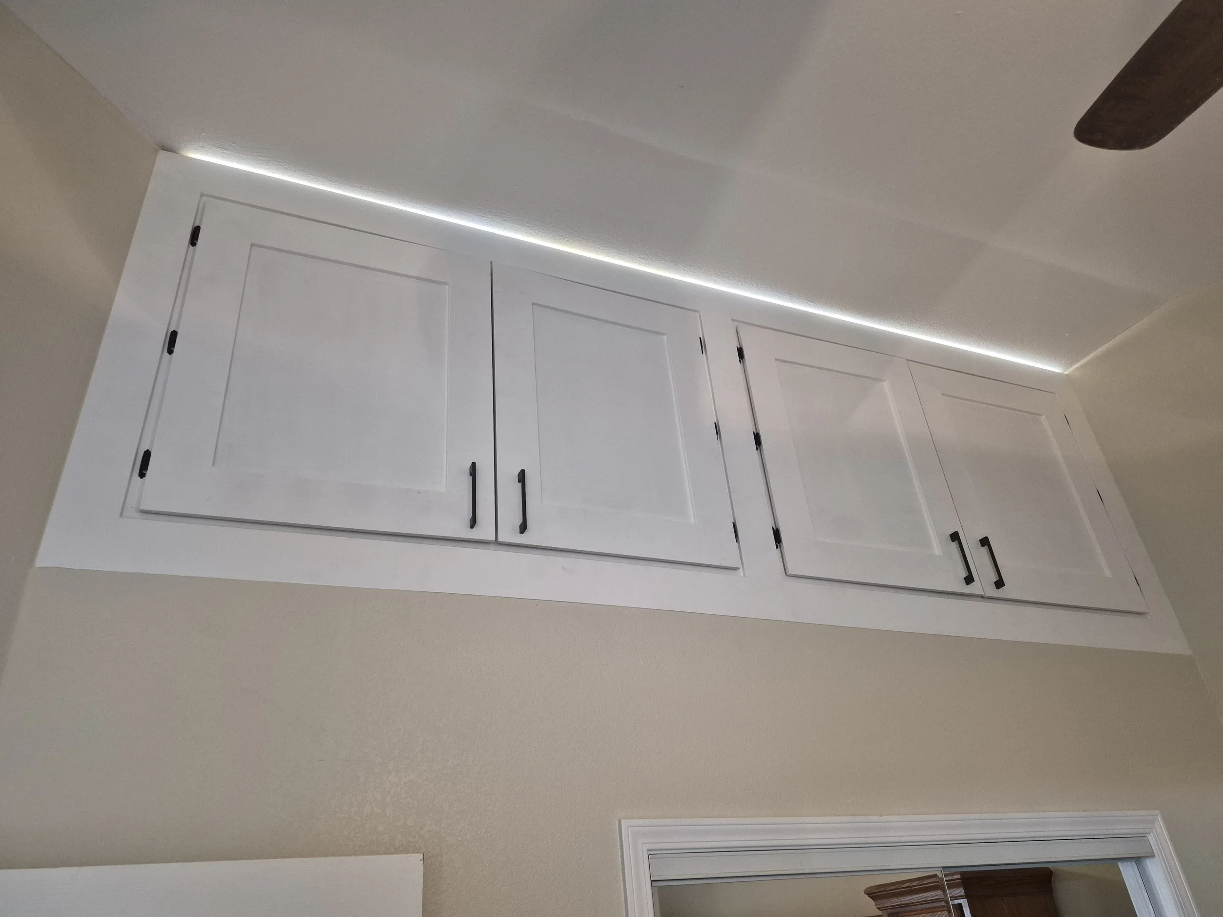 A ceiling with a recessed LED light strip along the edge, white cabinets with black handles, and a beige wall with a white vent.