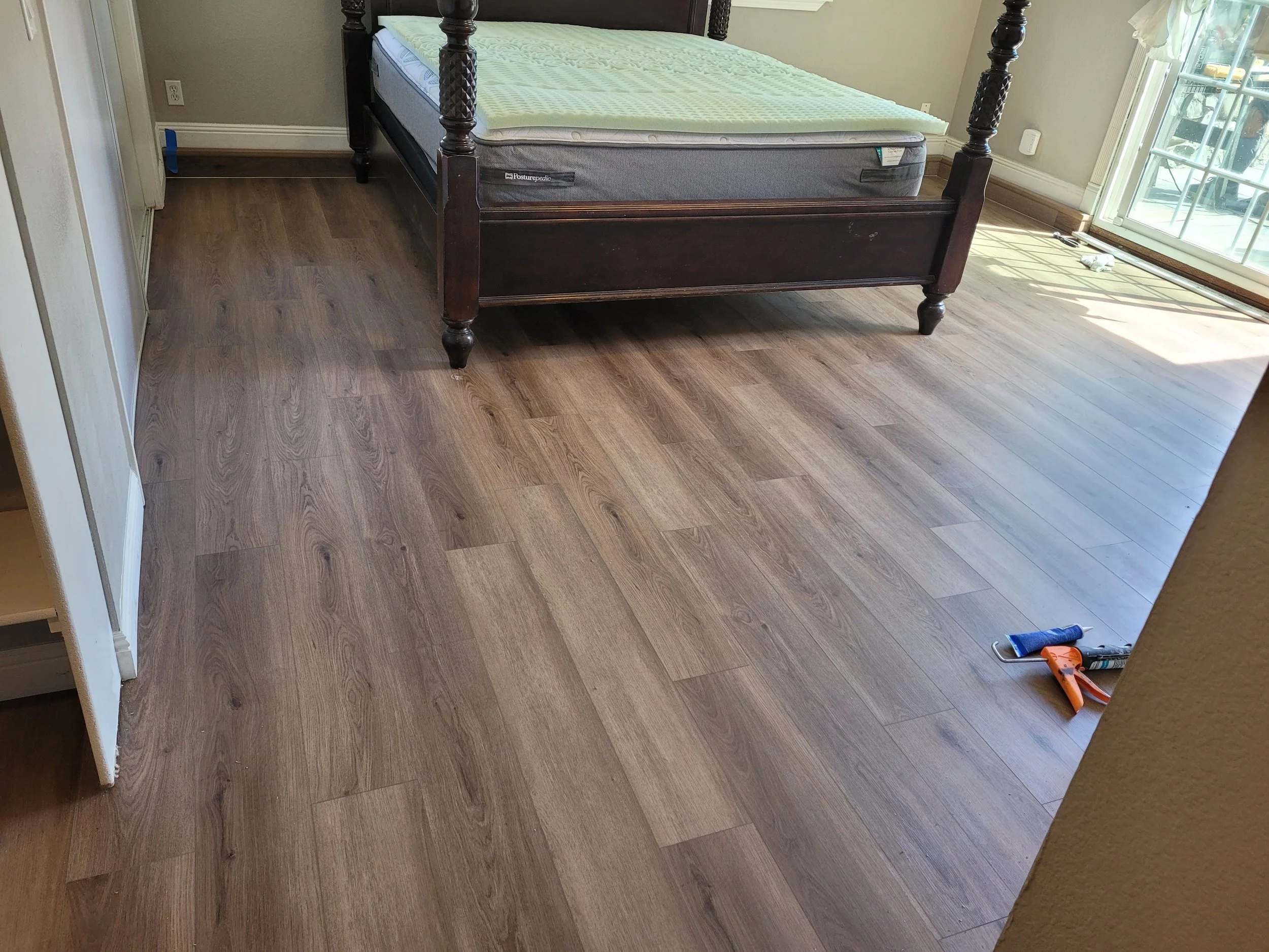 A bedroom with a wood floor, a dark wooden bed frame with a mattress, and tools on the floor near a sliding glass door.