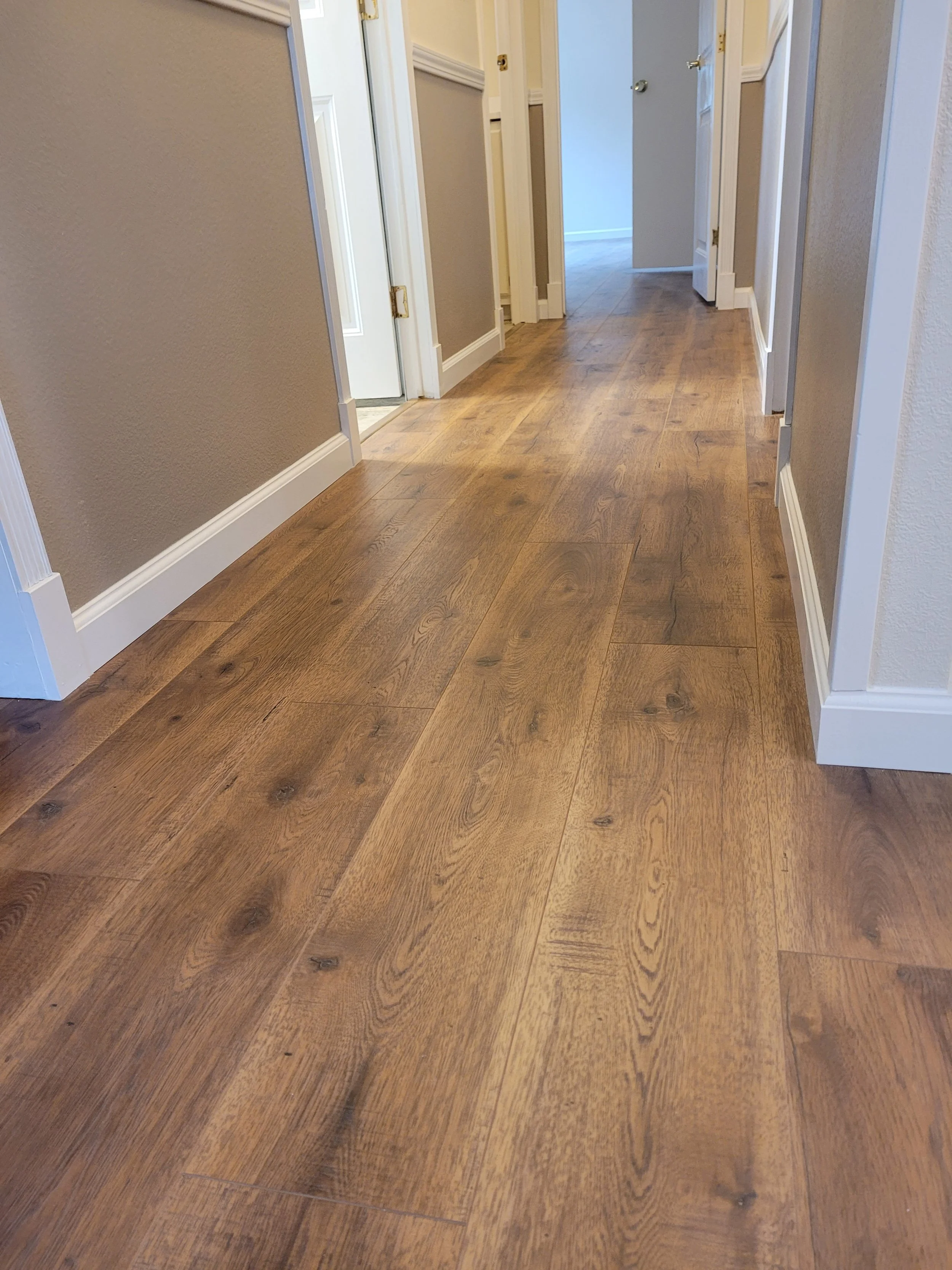 Wooden flooring in a hallway with several doorways, beige and white walls, and white baseboards.