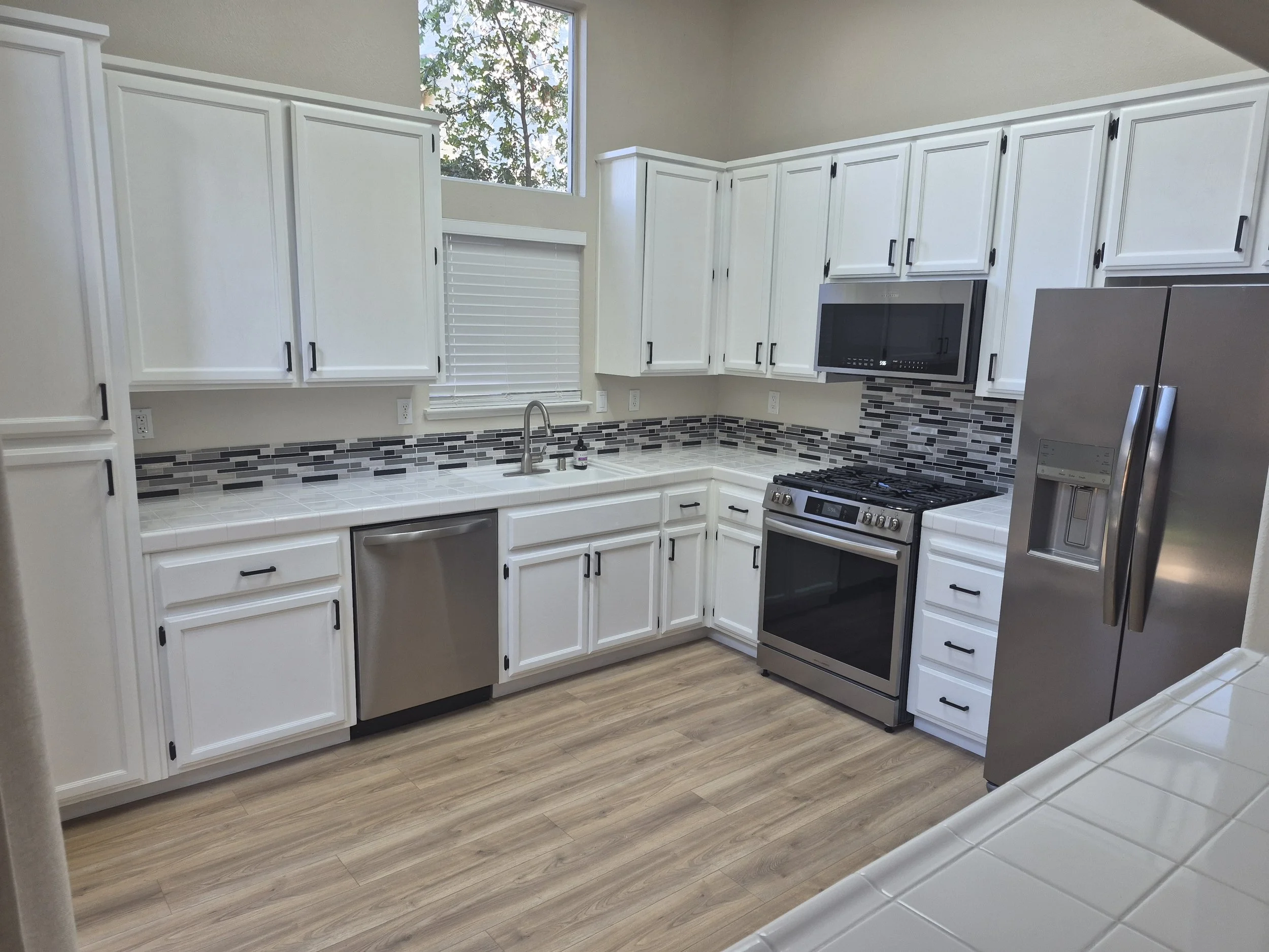 Modern kitchen with white cabinets, stainless steel appliances, gray and black tile backsplash, wooden flooring, and a window with trees outside.