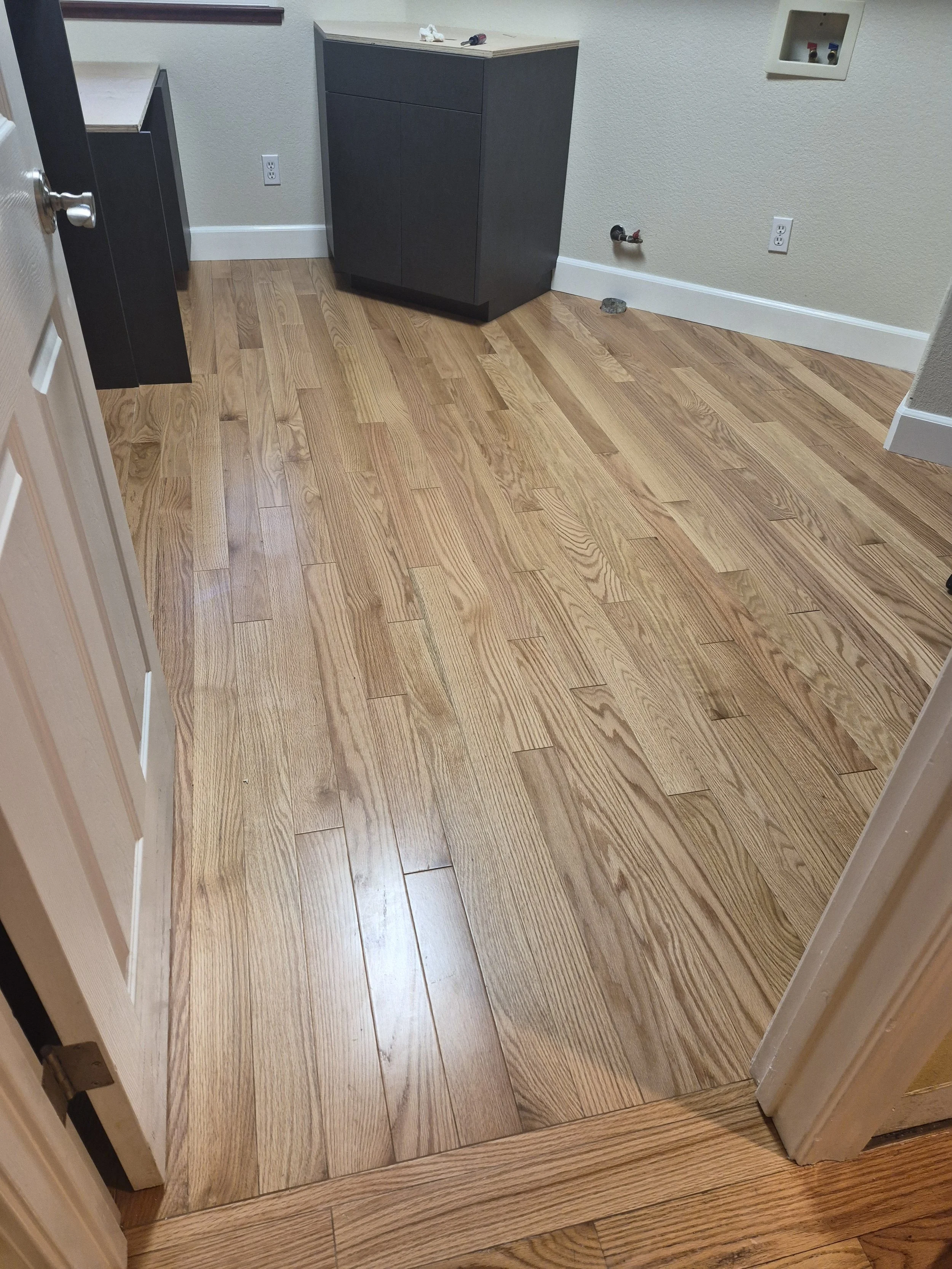 Empty room with wooden flooring, gray cabinets, electrical outlets, and plumbing fixtures.