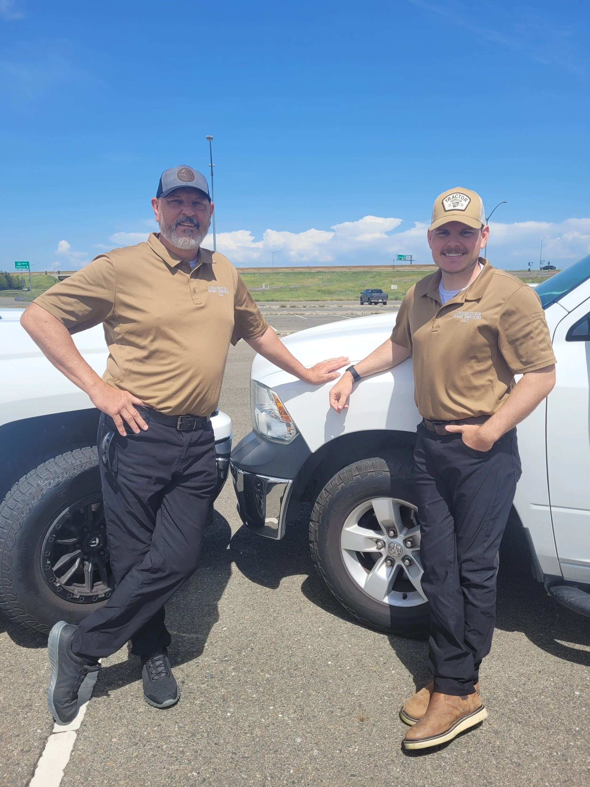 Two men in uniform standing beside white trucks on a highway. One man has a gray and black cap, the other a beige cap. They are smiling, with one resting a hand on the truck.