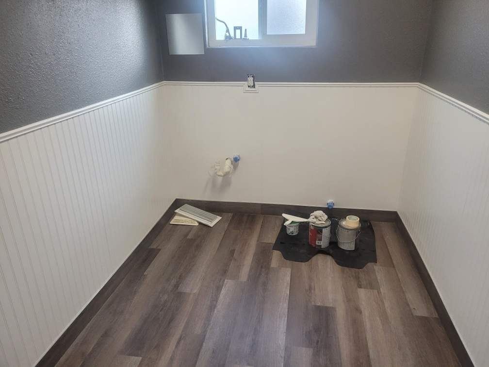 A small room under renovation, with half white beadboard wainscoting, dark gray upper walls, and a gray wood floor. There are paint supplies on the floor and an unfinished wall with a window.