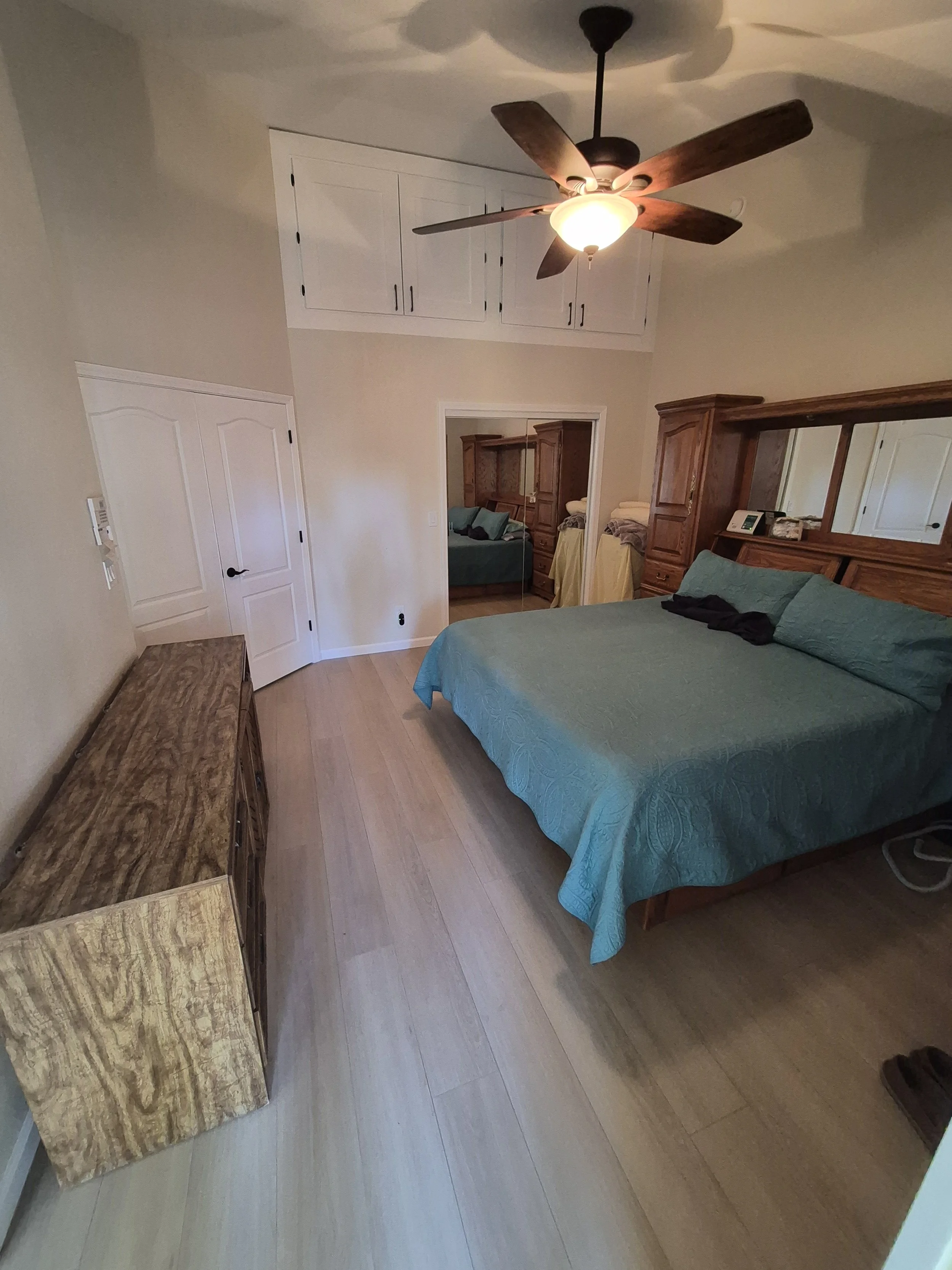 A bedroom with a ceiling fan, a bed with a turquoise quilt and pillows, a wooden dresser, and a mirror with built-in cabinets above the bed. There's a partially open door leading to another room with a couch and stored linens.