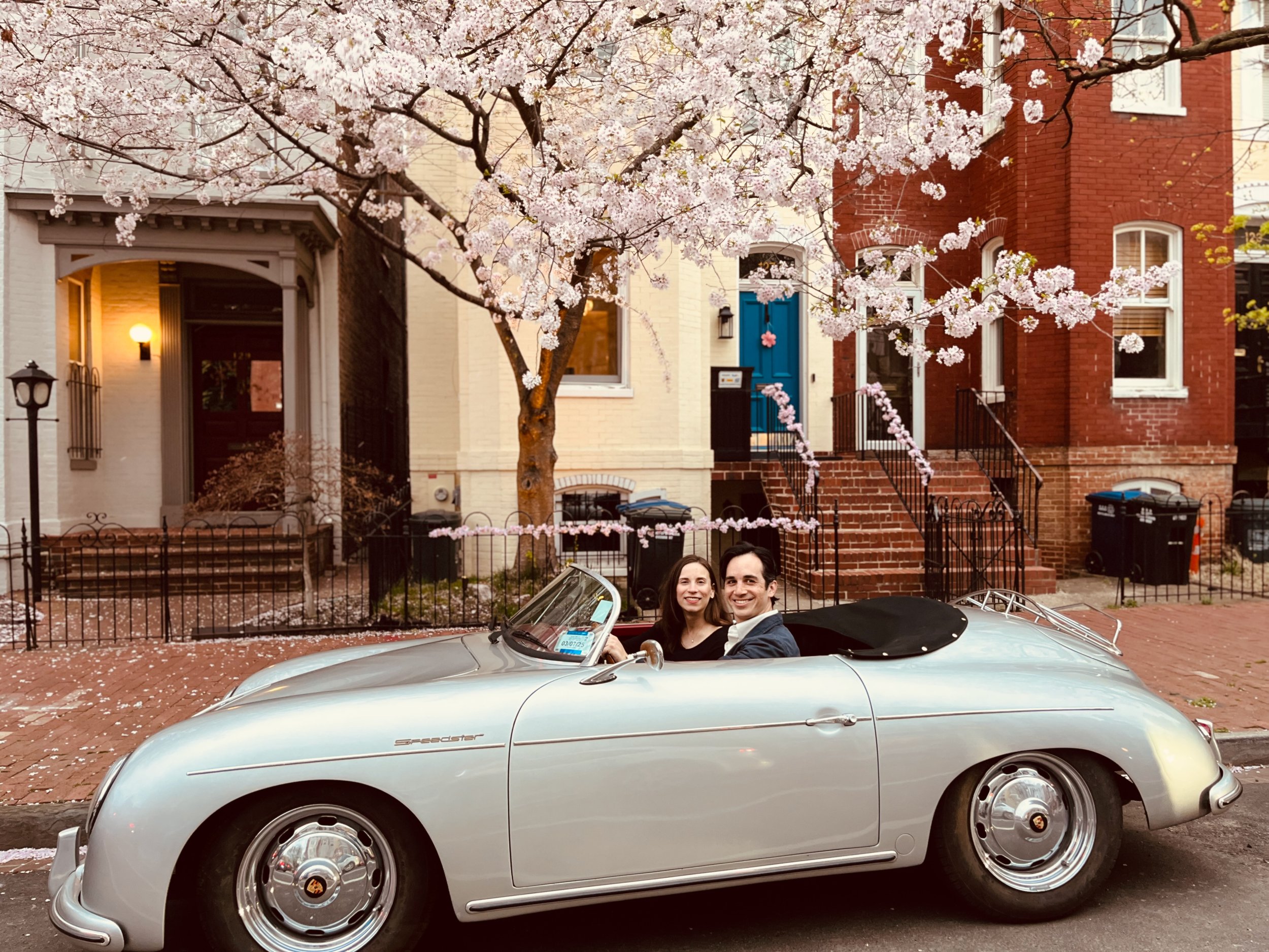 A couple seated in a vintage silver convertible car, parked on a street lined with cherry blossom trees and brick townhouses. The scene is serene, with blooming flowers creating a picturesque atmosphere.