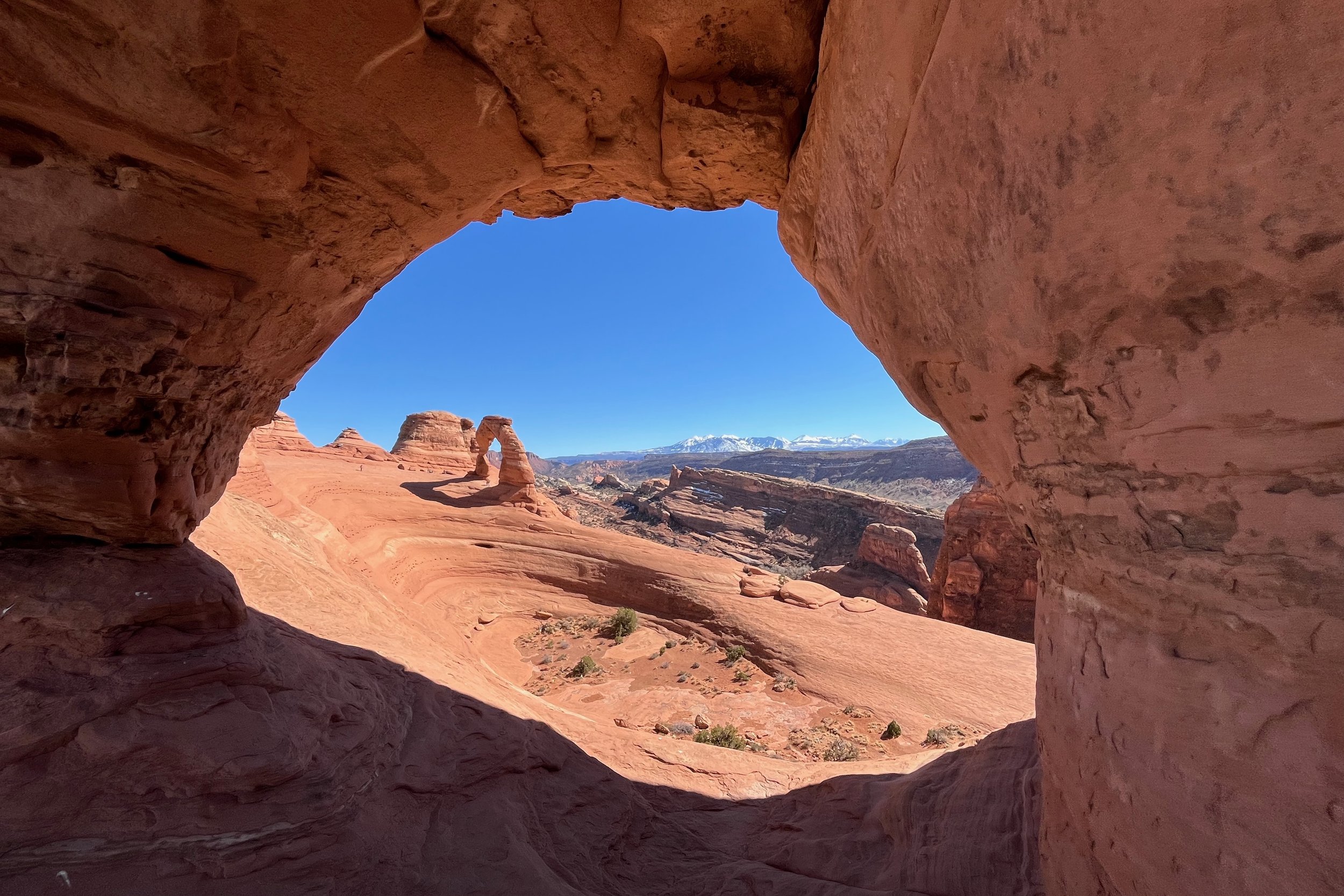 Delicate Arch viewed through a rocky cave opening, surrounded by red rock formations under a clear blue sky, at Arches National Park.