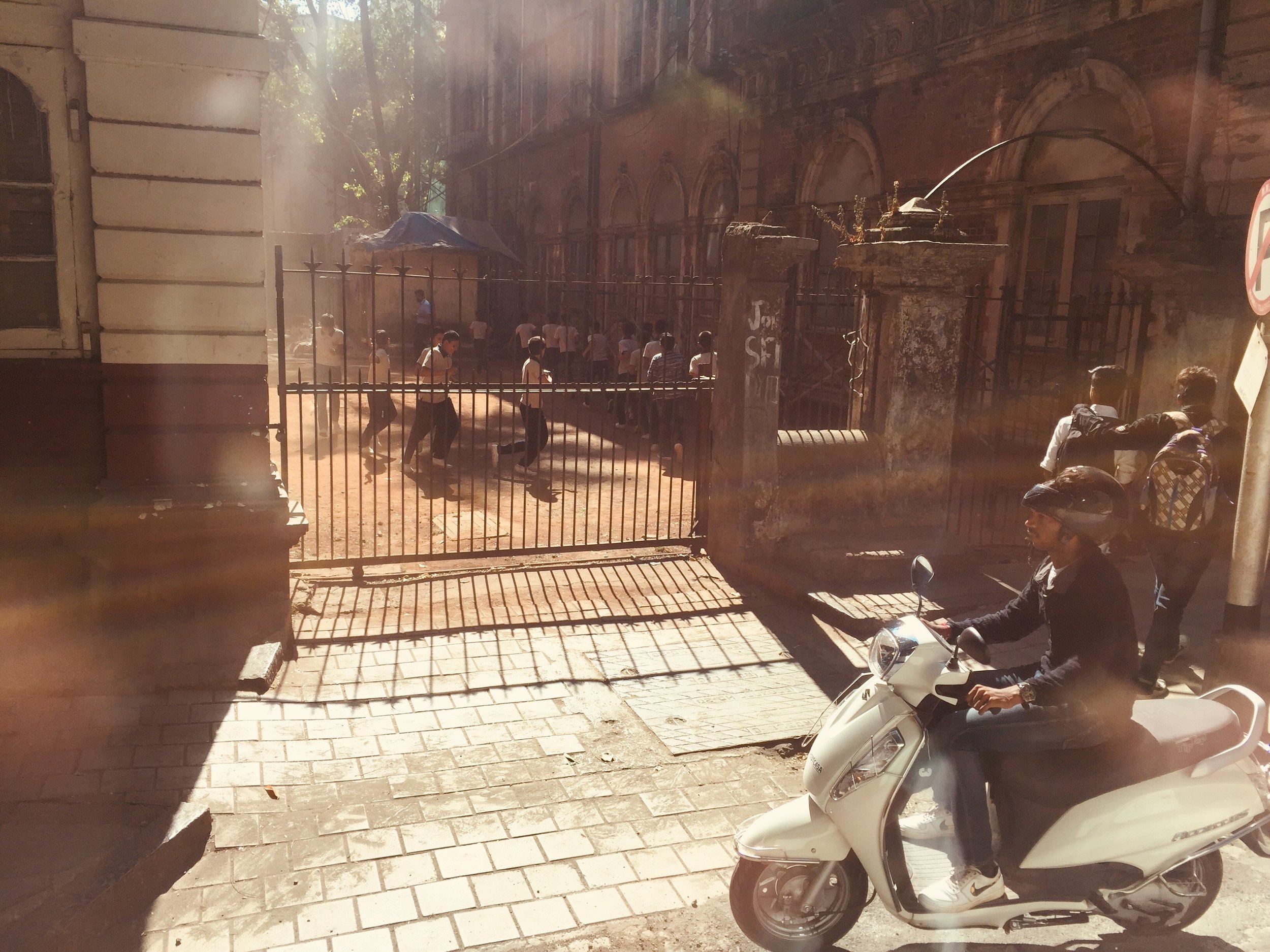 Person riding a scooter on a sunlit street next to a building and a fenced area with people inside.