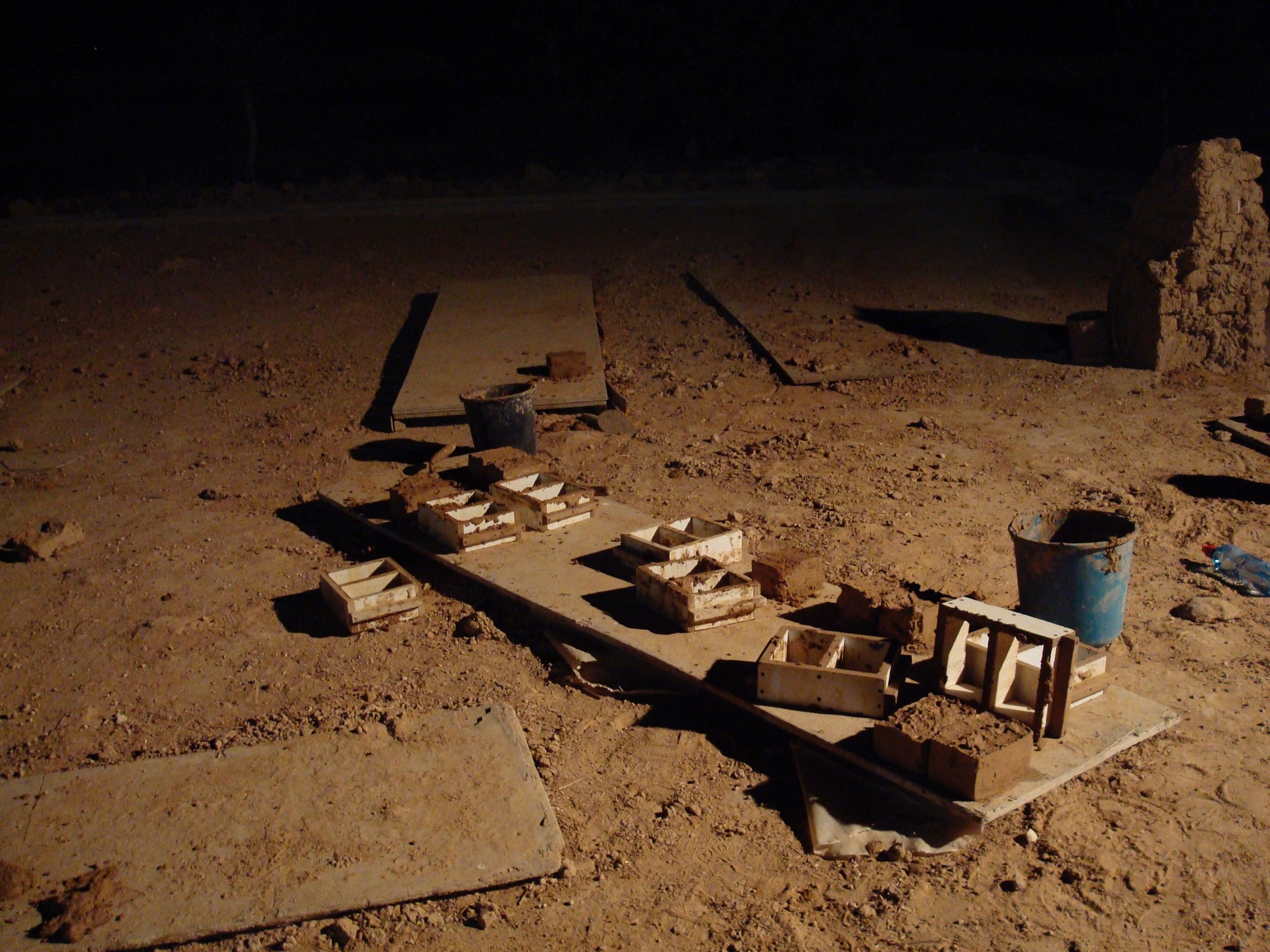 Outdoor scene with several wooden molds filled with wet clay on the ground, possibly for brick making, alongside a blue bucket and dirt in the background.