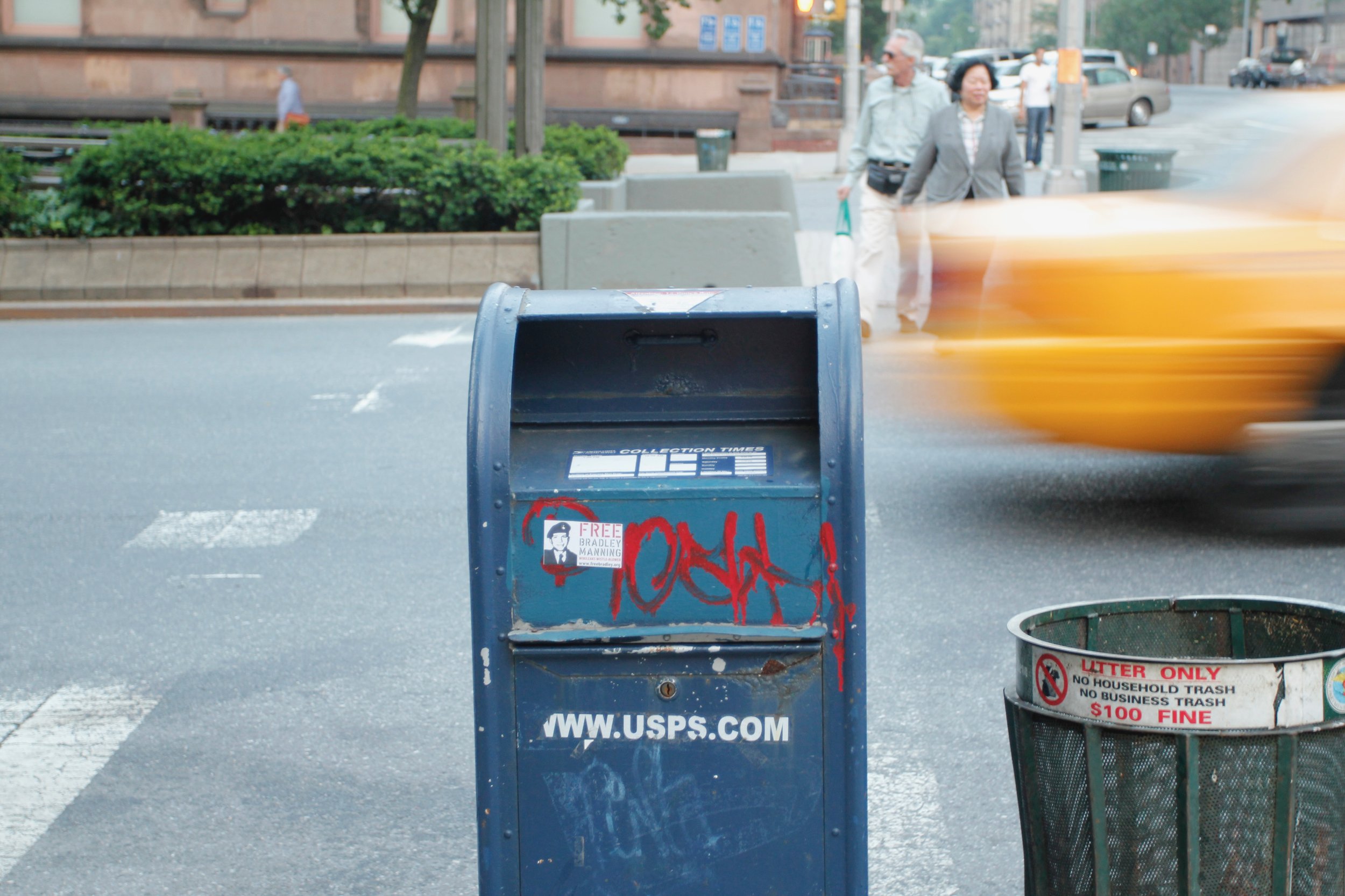 USPS mailbox with graffiti, trash can nearby, blurred yellow taxi, people walking on sidewalk, urban street scene.