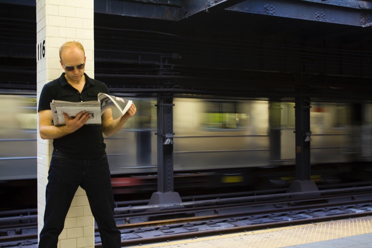 Man wearing sunglasses reading a newspaper on a subway platform with a moving train in the background.