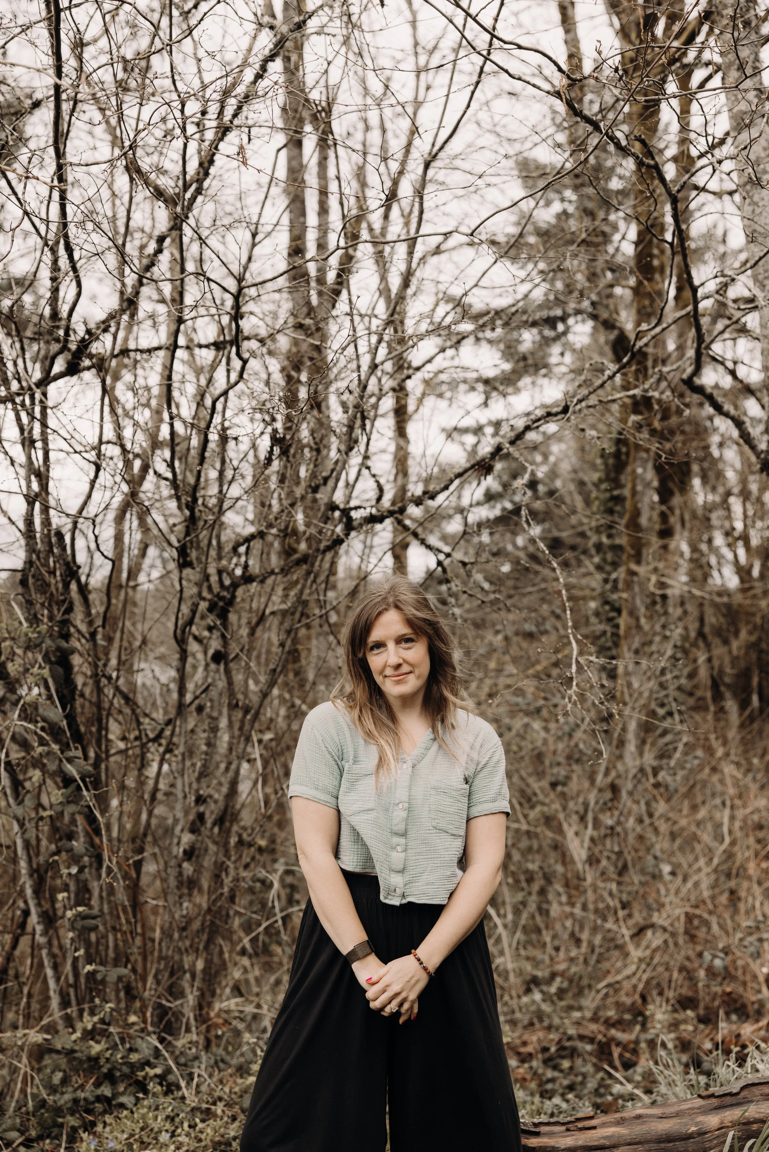 Laura, a Portland Doula, is wearing a light green top and black pants. Laura stands in a leafless forest, looking at the camera with a slight smile. The background is filled with bare trees and tangled branches, creating a serene, natural setting.