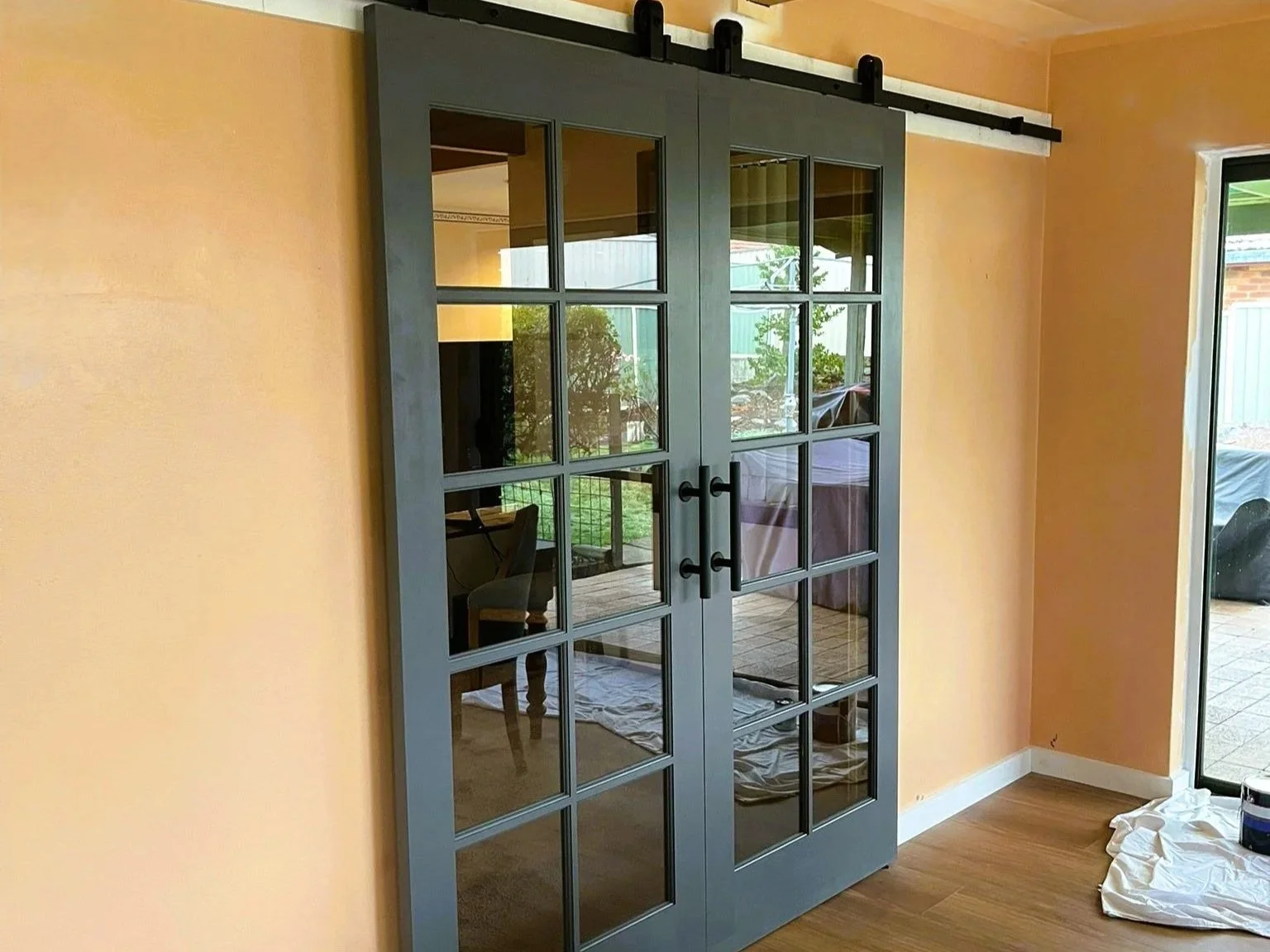 Interior of a room with a gray sliding barn door with glass panes, a peach-colored wall, a sliding glass door leading outside, and some painting supplies on the floor.