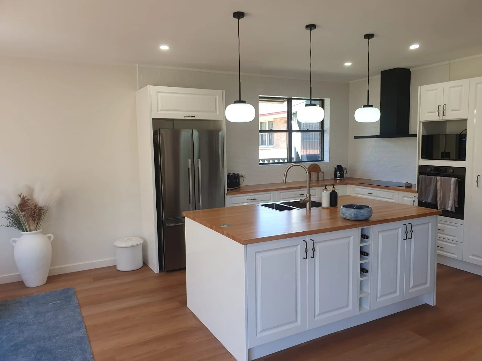 Modern kitchen with white cabinets, wooden countertops, stainless steel refrigerator, built-in oven, black range hood, island with sink, pendant lights, window with view of neighboring house, minimal decor.