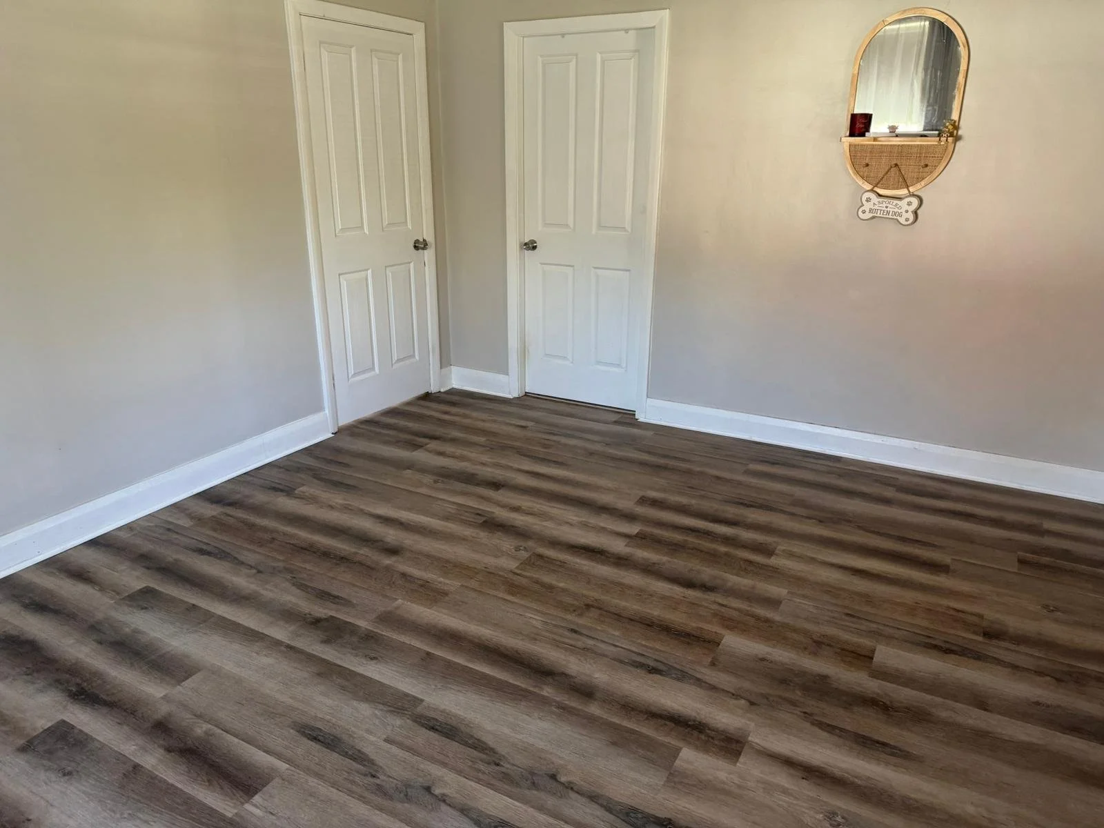 Empty room with light beige walls, white baseboards, two white doors, a mirror on the wall, and wood laminate flooring.