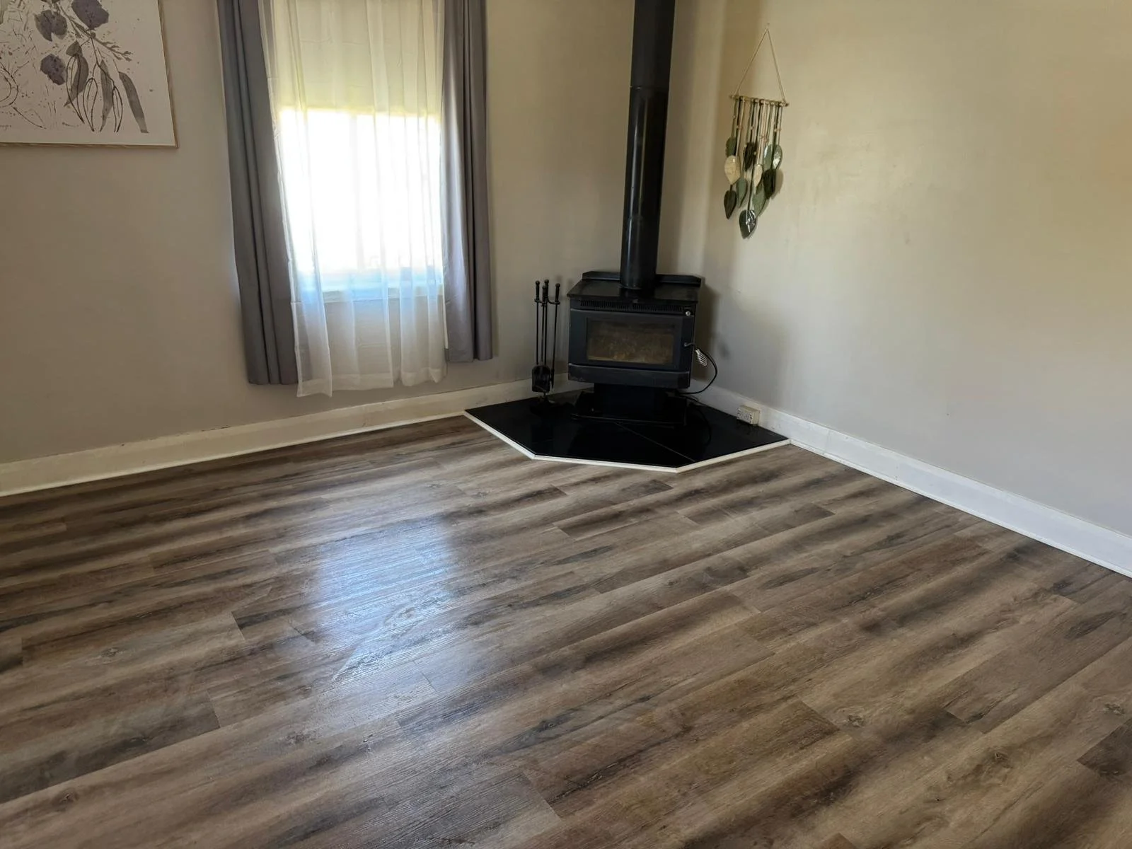Empty living room with wooden floor, window with curtains, and a black wood stove in the corner.