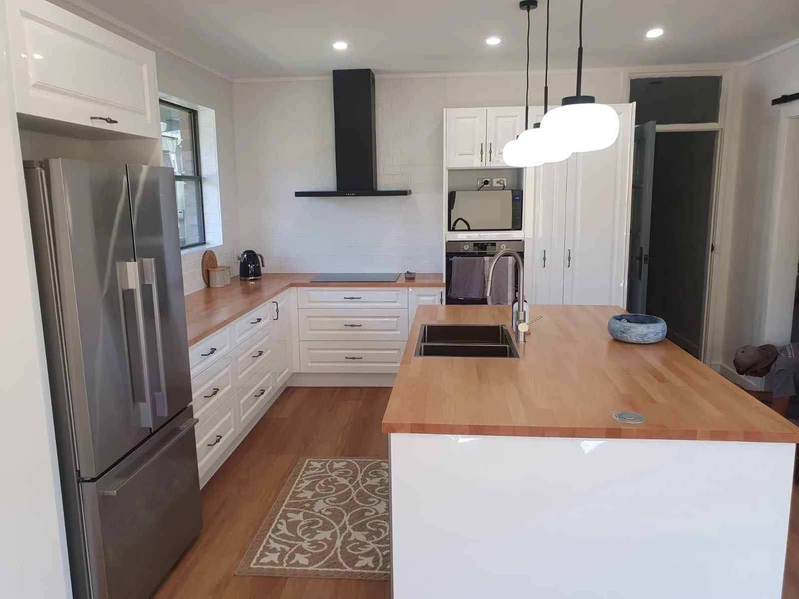 Modern kitchen with white cabinets, wooden countertops, stainless steel refrigerator, a kitchen island with a sink, pendant lighting, a window, and a person partially visible on the right.