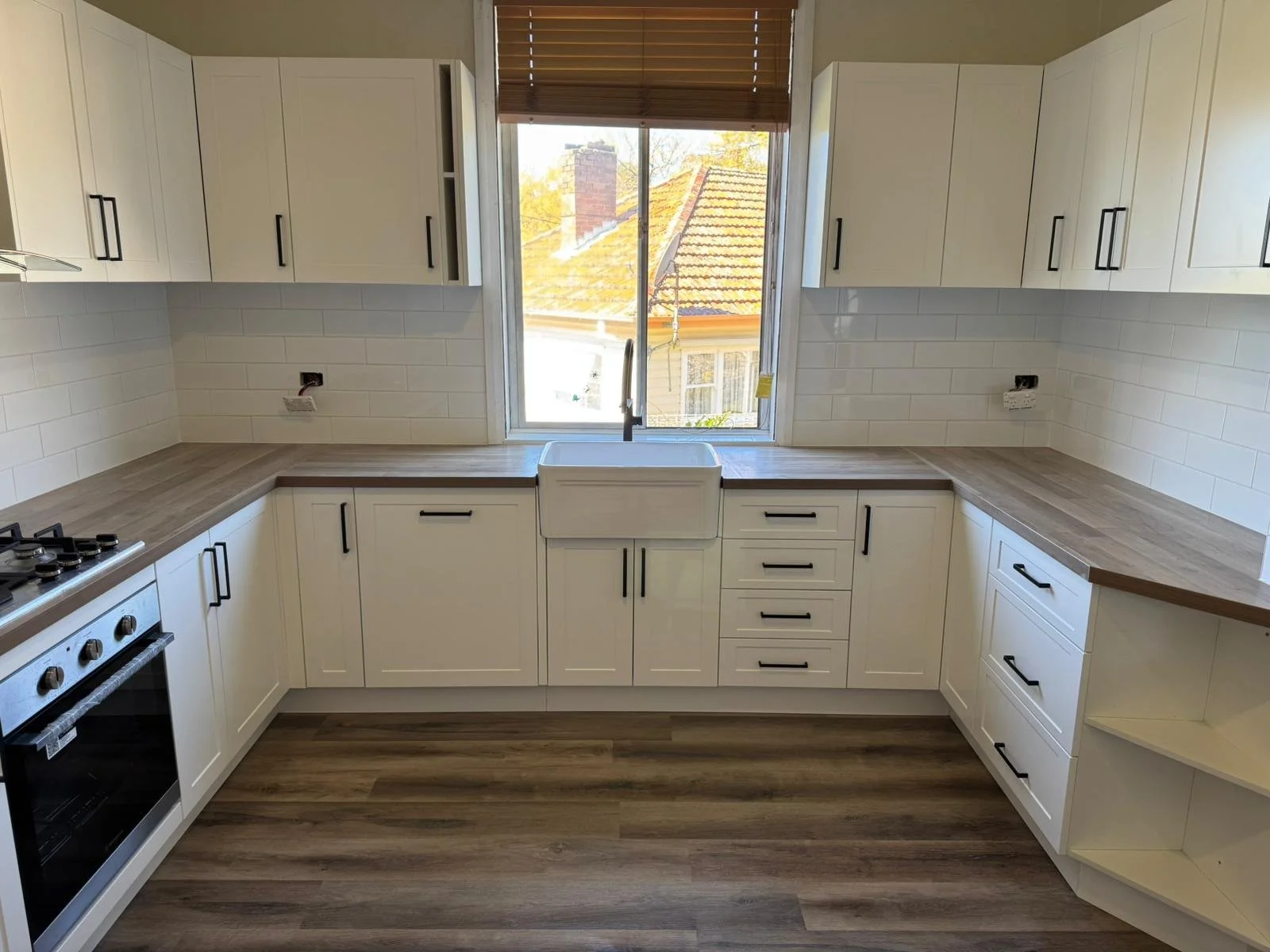 Kitchen with white cabinets, wood countertops, a window with blinds, a sink under the window, and a stove on the left. The kitchen is bright with natural light and has wood flooring.