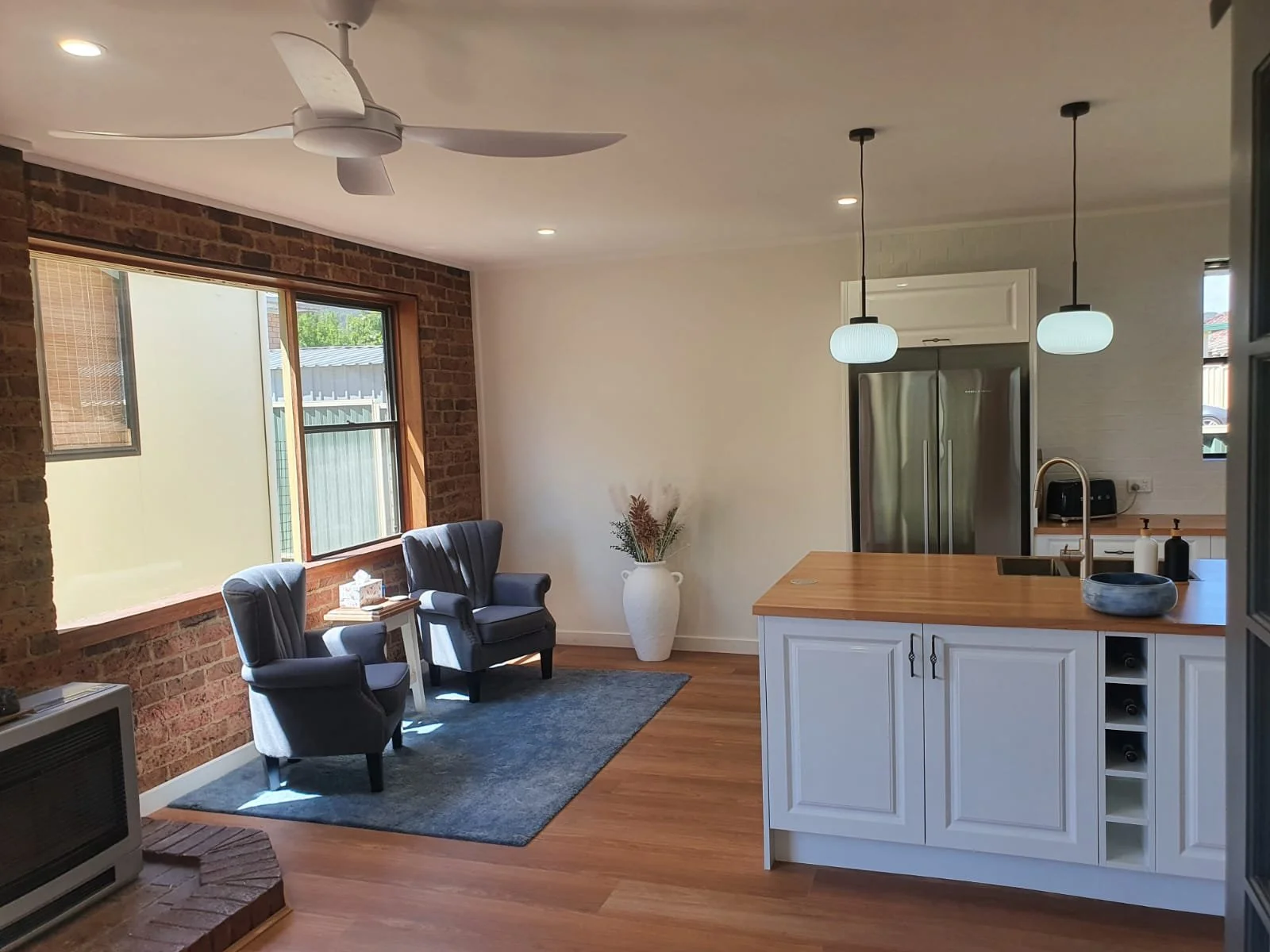 Kitchen and living room with brick wall, large window, two gray armchairs, white vase with dried flowers, wooden countertops, white cabinets, stainless steel refrigerator, ceiling fan, and pendant lights.