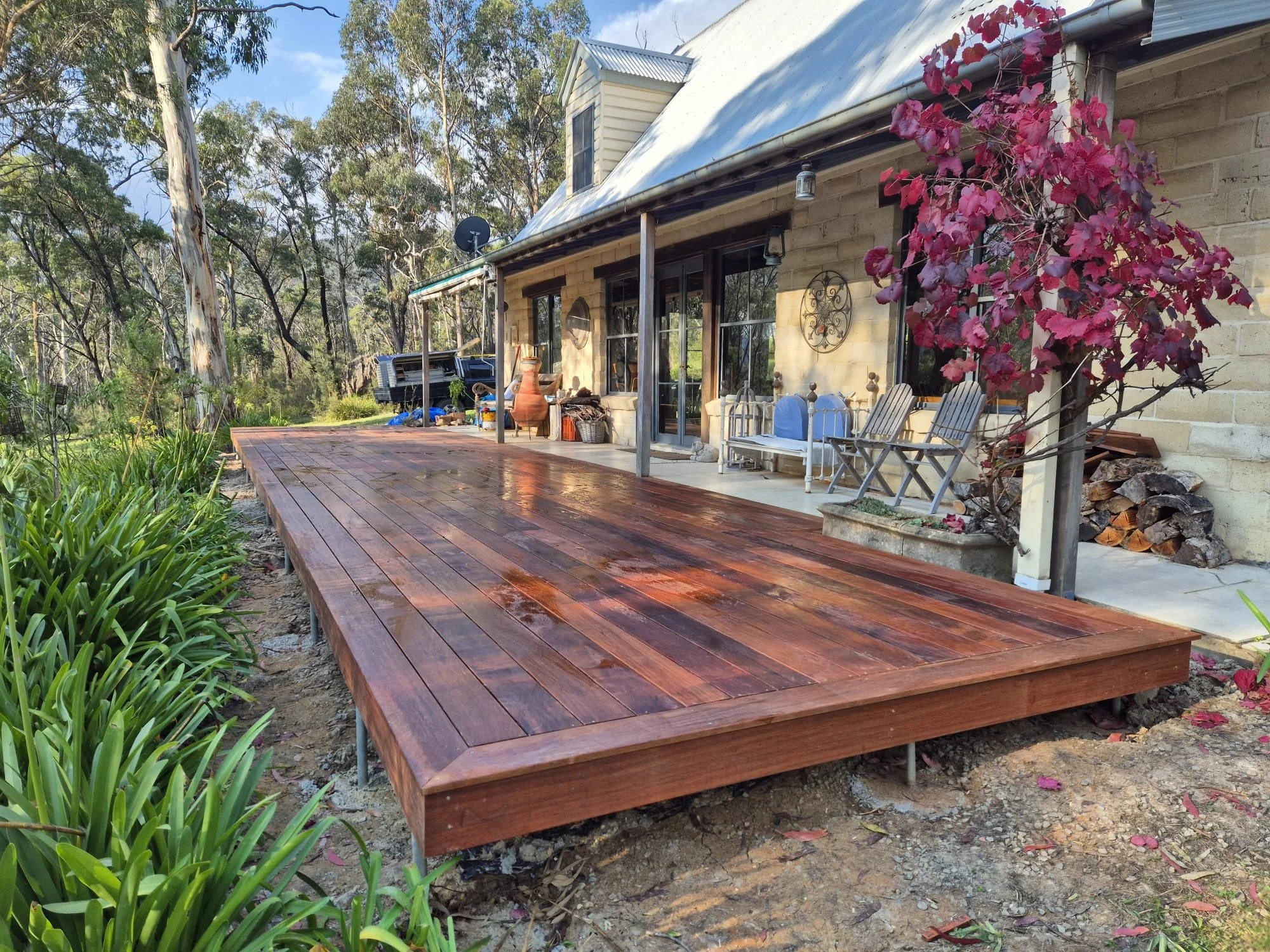 A wooden deck attached to the back of a house with outdoor furniture, potted plants, and a firepit, surrounded by trees and greenery.