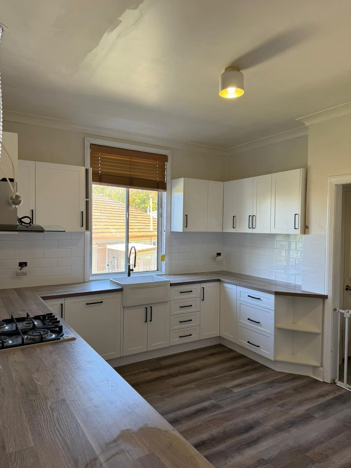 Empty kitchen with white cabinets, wooden countertops, a window with blinds, and a stove, with a ceiling light turned on.