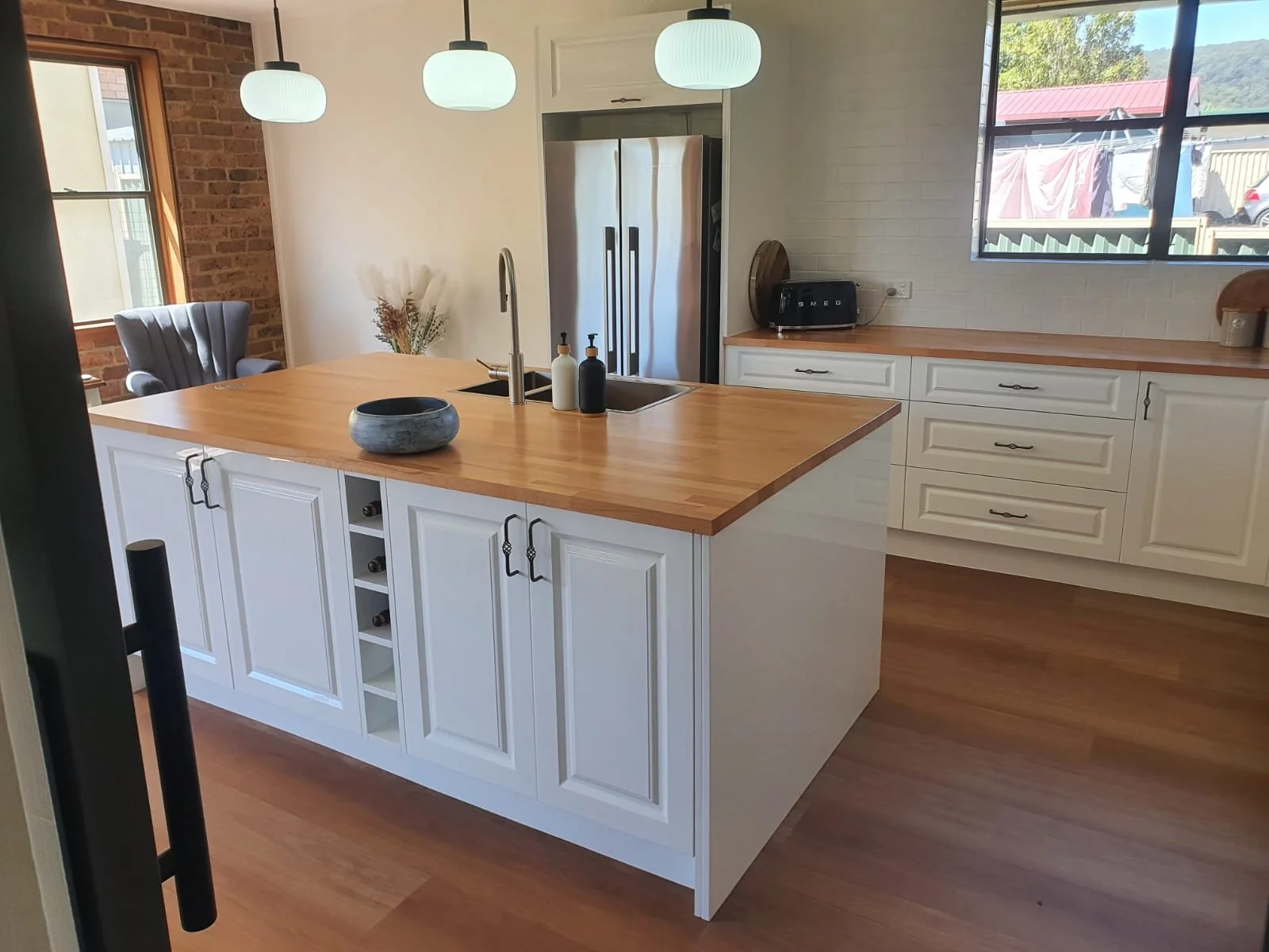 Modern kitchen with a large wooden island, white cabinets, stainless steel refrigerator, and hanging pendant lights. A window reveals a backyard with laundry hanging on a clothesline.