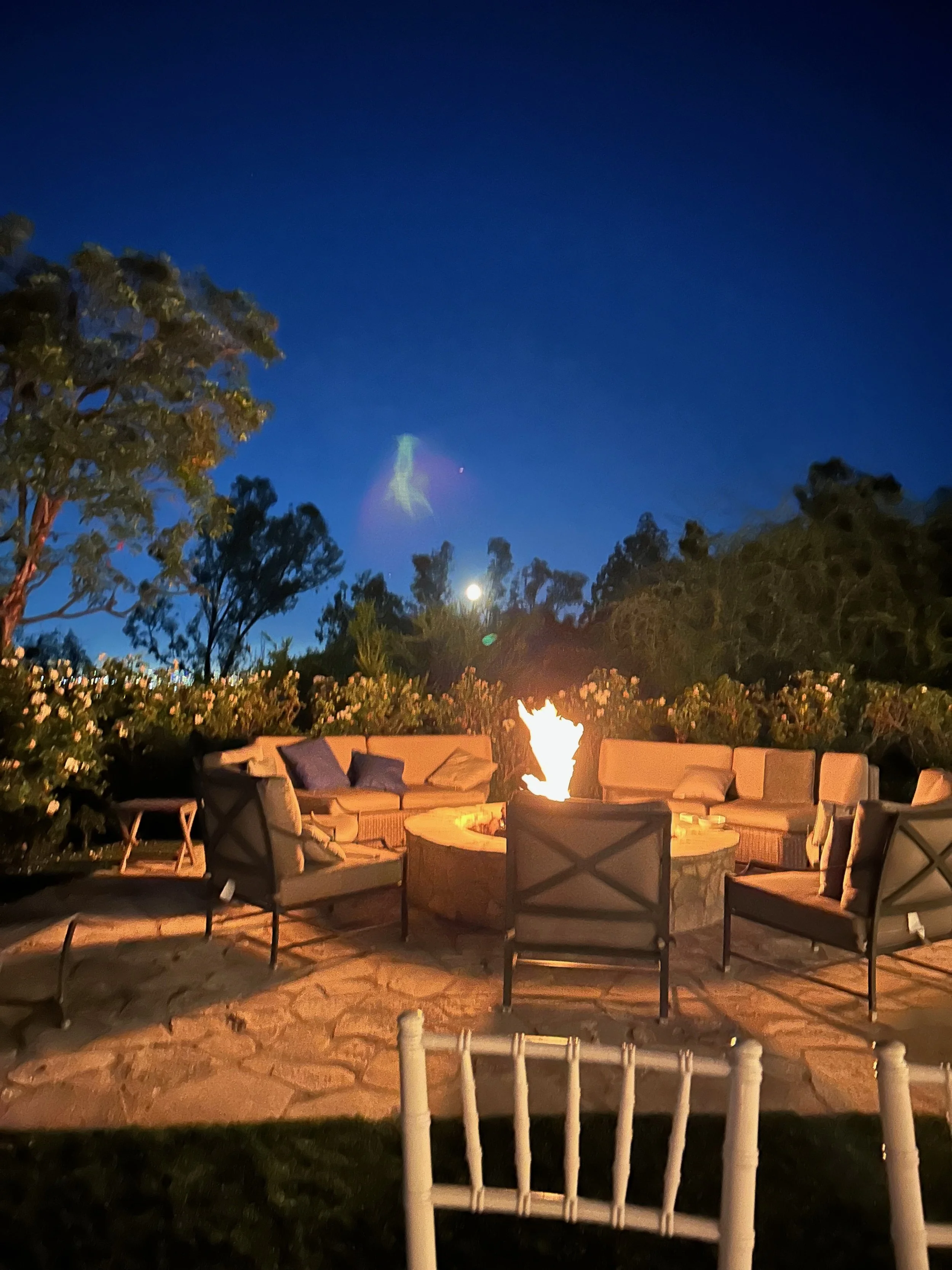 Outdoor patio with fire pit surrounded by cushioned chairs and sofas, set against a night sky with trees in the background signifying supernatural visits.