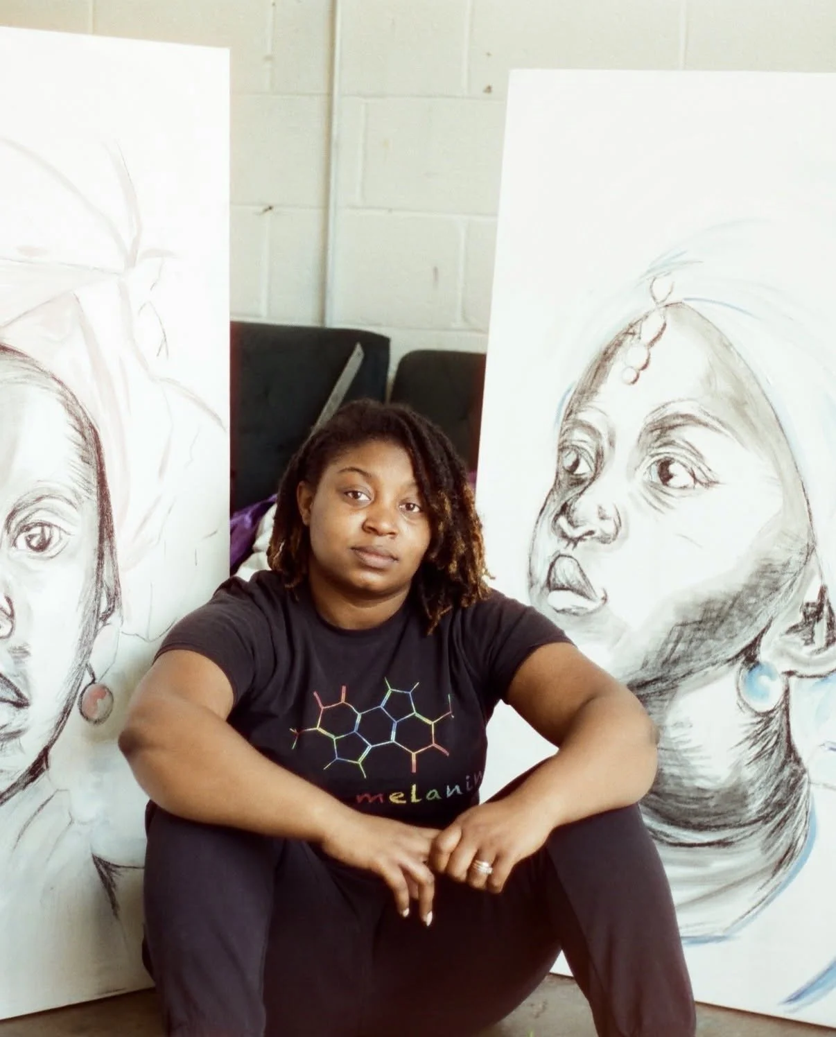 A woman with shoulder-length curly hair sitting in front of large canvas sketches of faces, inside a room with white brick wall.