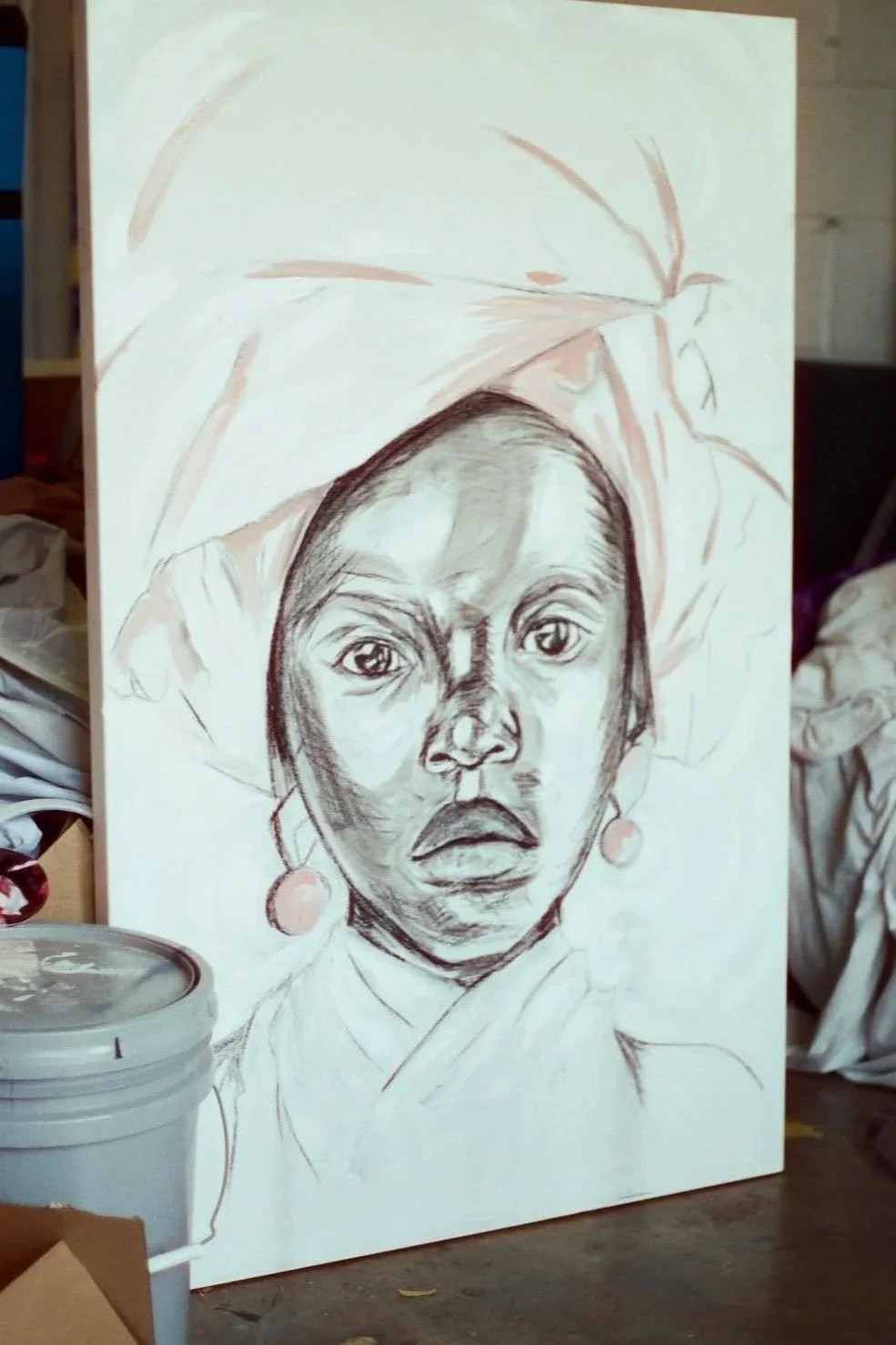 A black and white sketch of a woman wearing a headscarf and earrings, with expressive facial features, in progress, on a white canvas in an art studio.