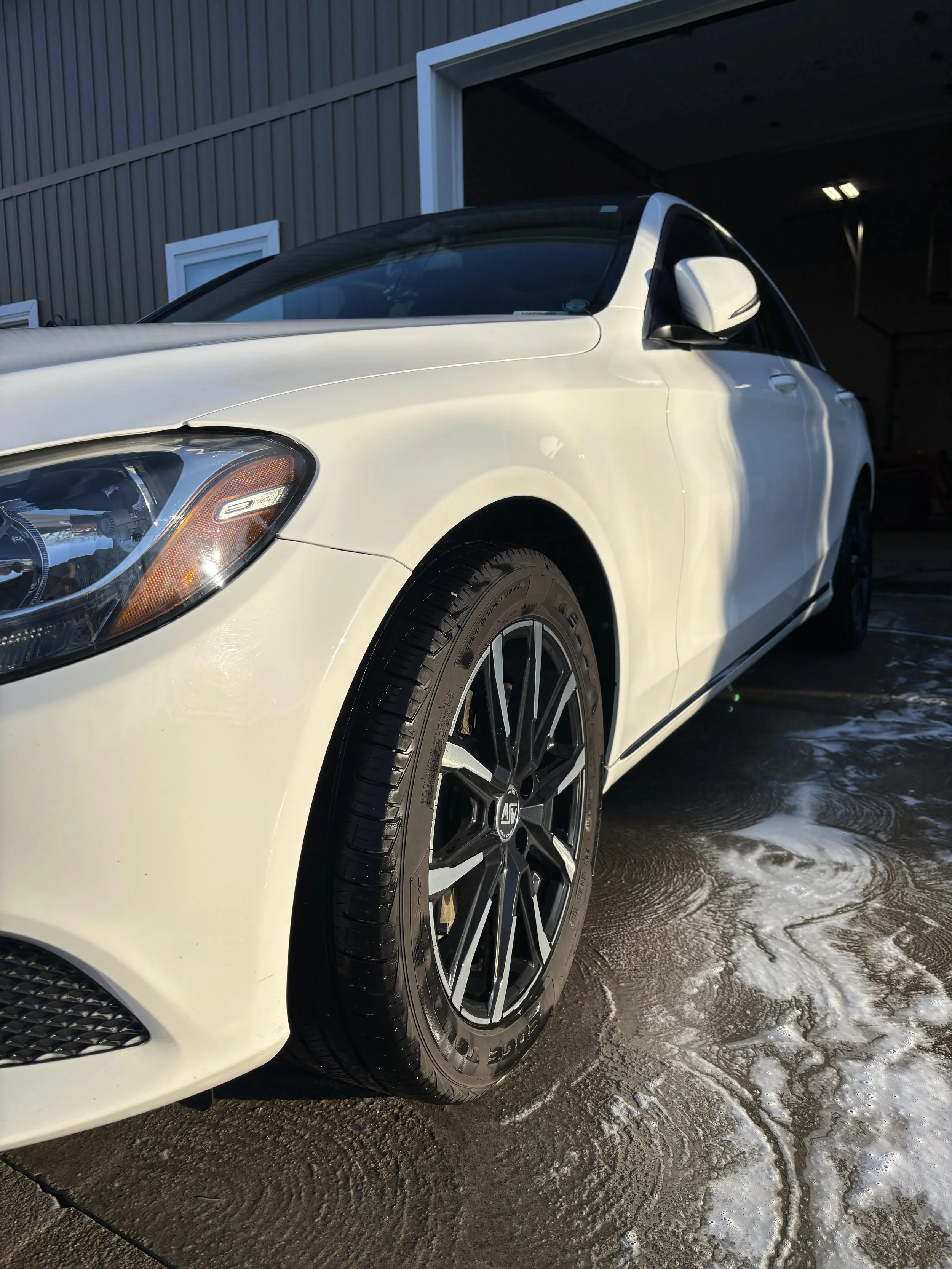 A white sedan car parked outside in front of a garage, with sunlight reflecting off the hood and tire, and soap suds and water on the ground.
