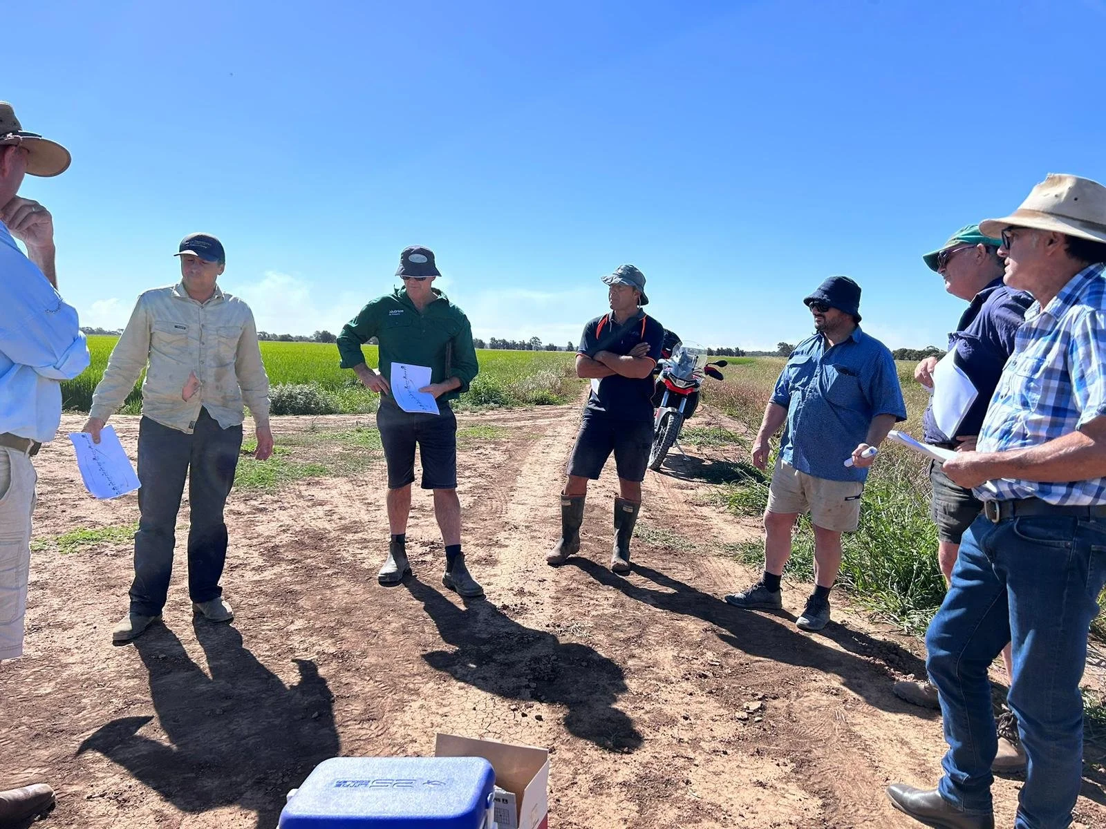 Group of people standing in a field having a discussion, some are holding papers, one has a motorcycle in the background, and the weather is sunny and clear.